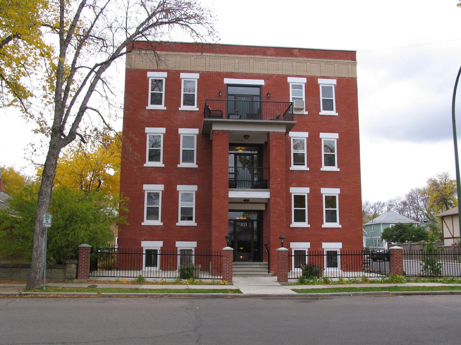 A red brick multi-story apartment building with three balconies and white window frames, surrounded by a black fence, with trees and other houses in the background.