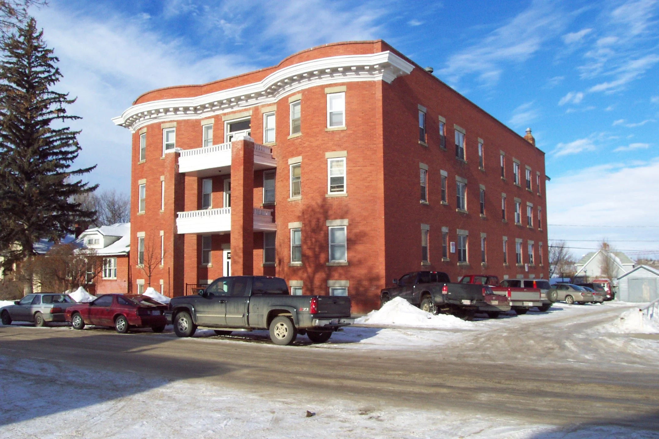A brick four-story apartment building with white decorative trim under a blue partly cloudy sky. Several cars parked along the street in front, with snow on the ground.
