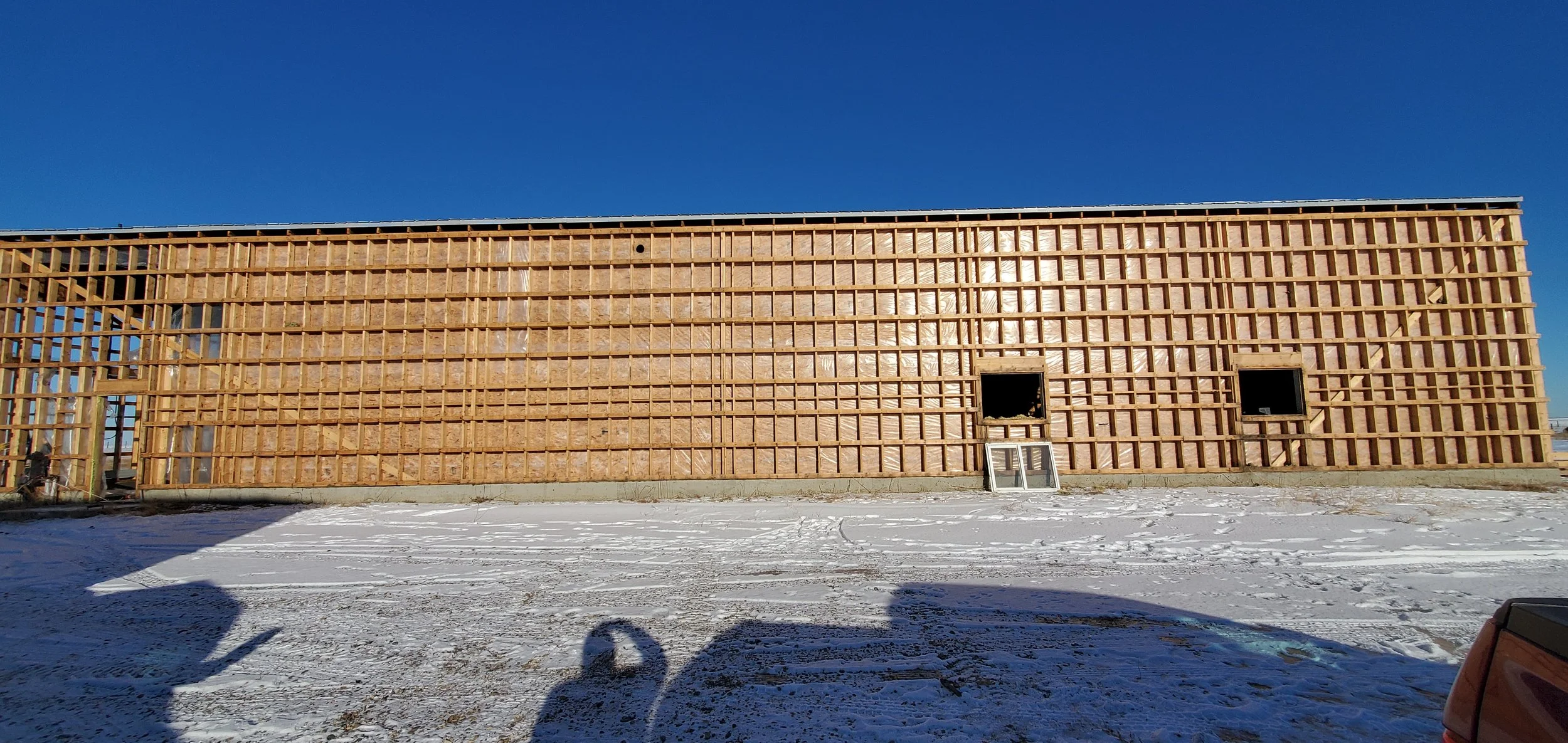 Wooden framing of a building under construction against a clear blue sky and snow-covered ground.
