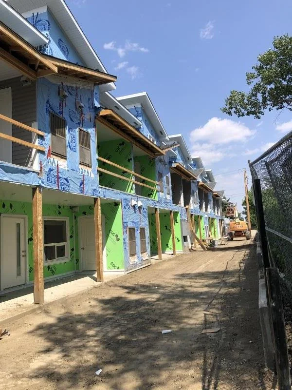 Row of attached new townhouses under construction, with green and blue insulation wrap, wooden supports, and construction equipment in the background, on a dirt road with trees against a blue sky.