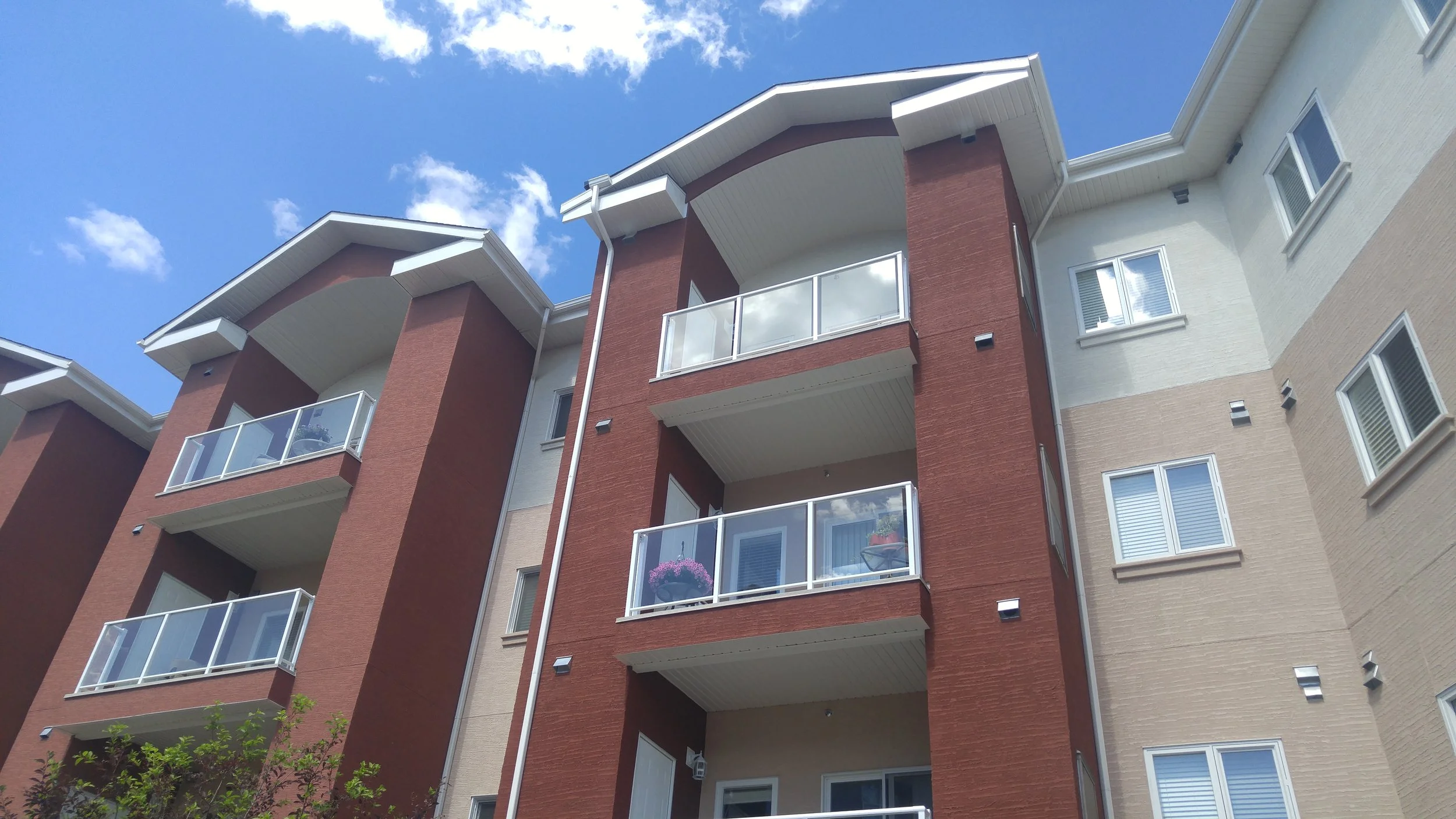 Low-angle view of a modern multi-story apartment building with balconies, white and red brick exterior, and a blue sky with clouds in the background.