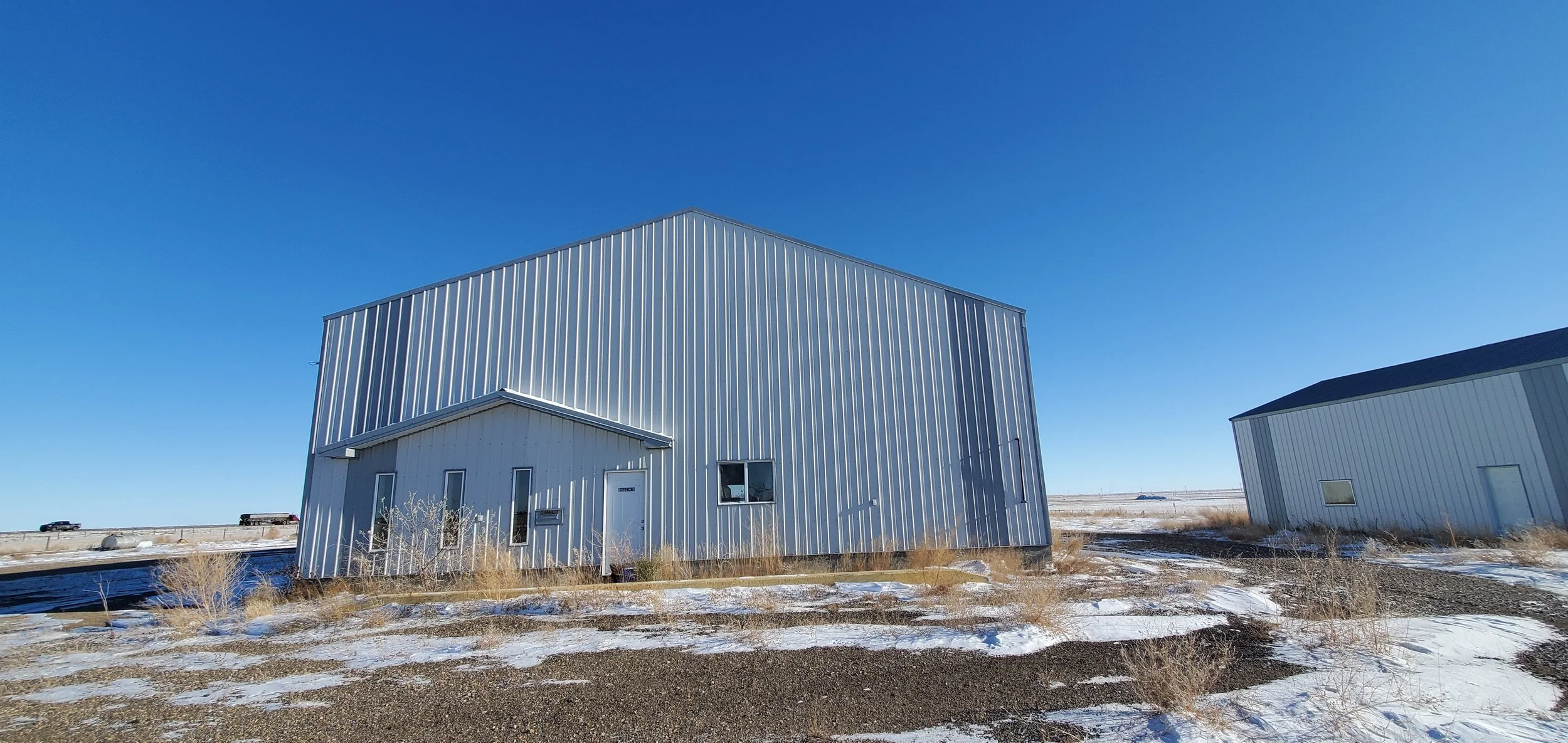 A large metal building with a sloped roof in a landscape with snow, dry grass, and a clear blue sky.
