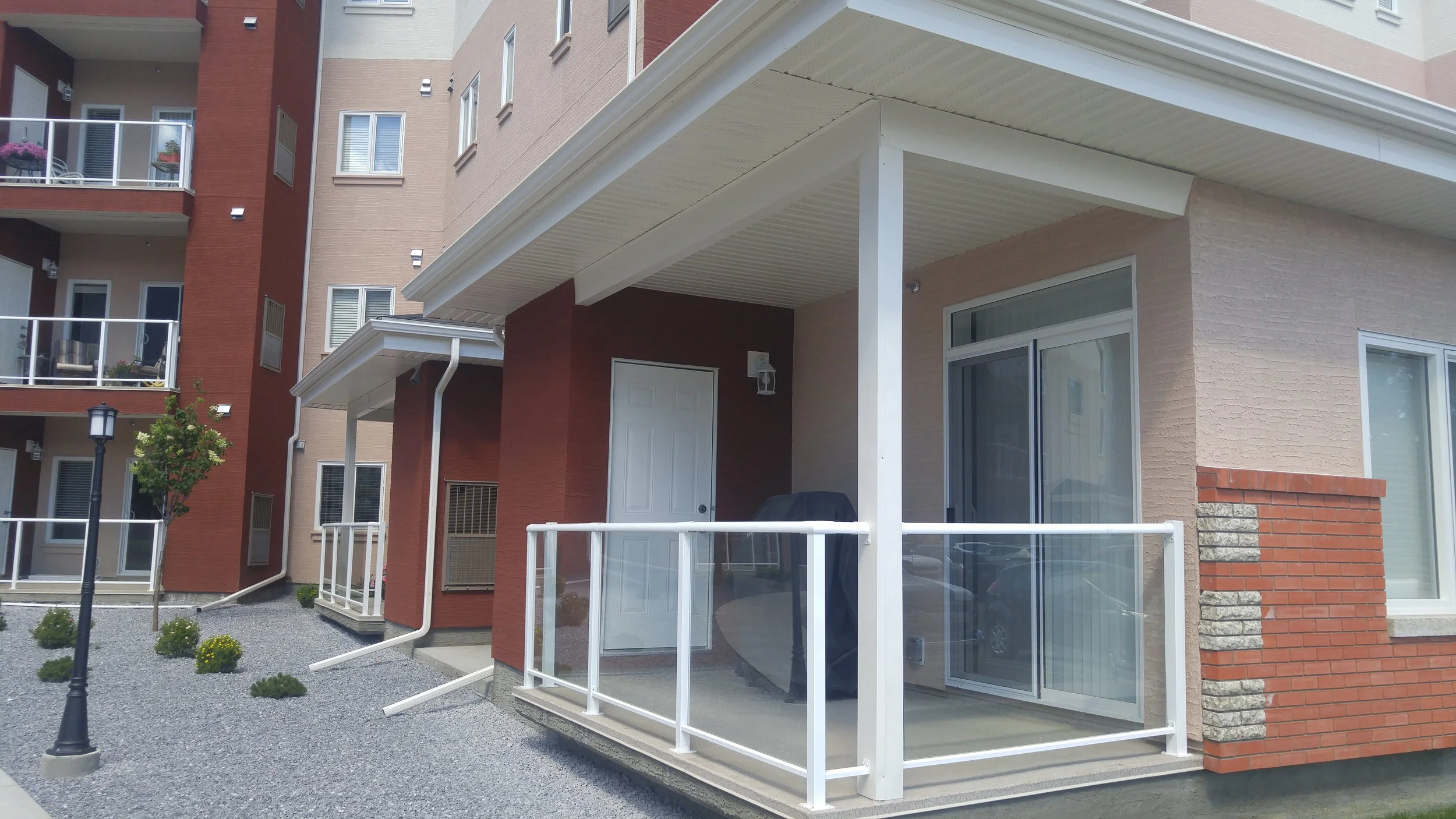 Exterior view of a modern multi-story apartment building with balconies, windows, and a small porch area with sliding glass doors, white railing, and a gravel walkway.