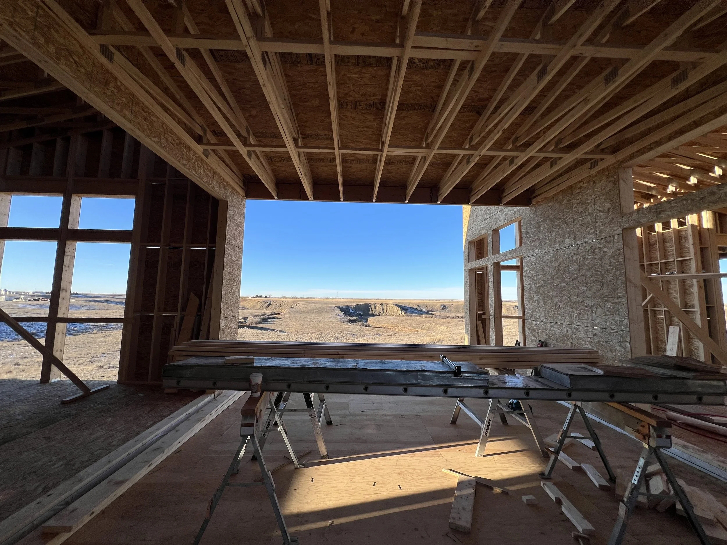 Interior view of a house under construction showing exposed wooden framing, OSB sheathing on walls, and a view of the outdoors with clear blue sky.