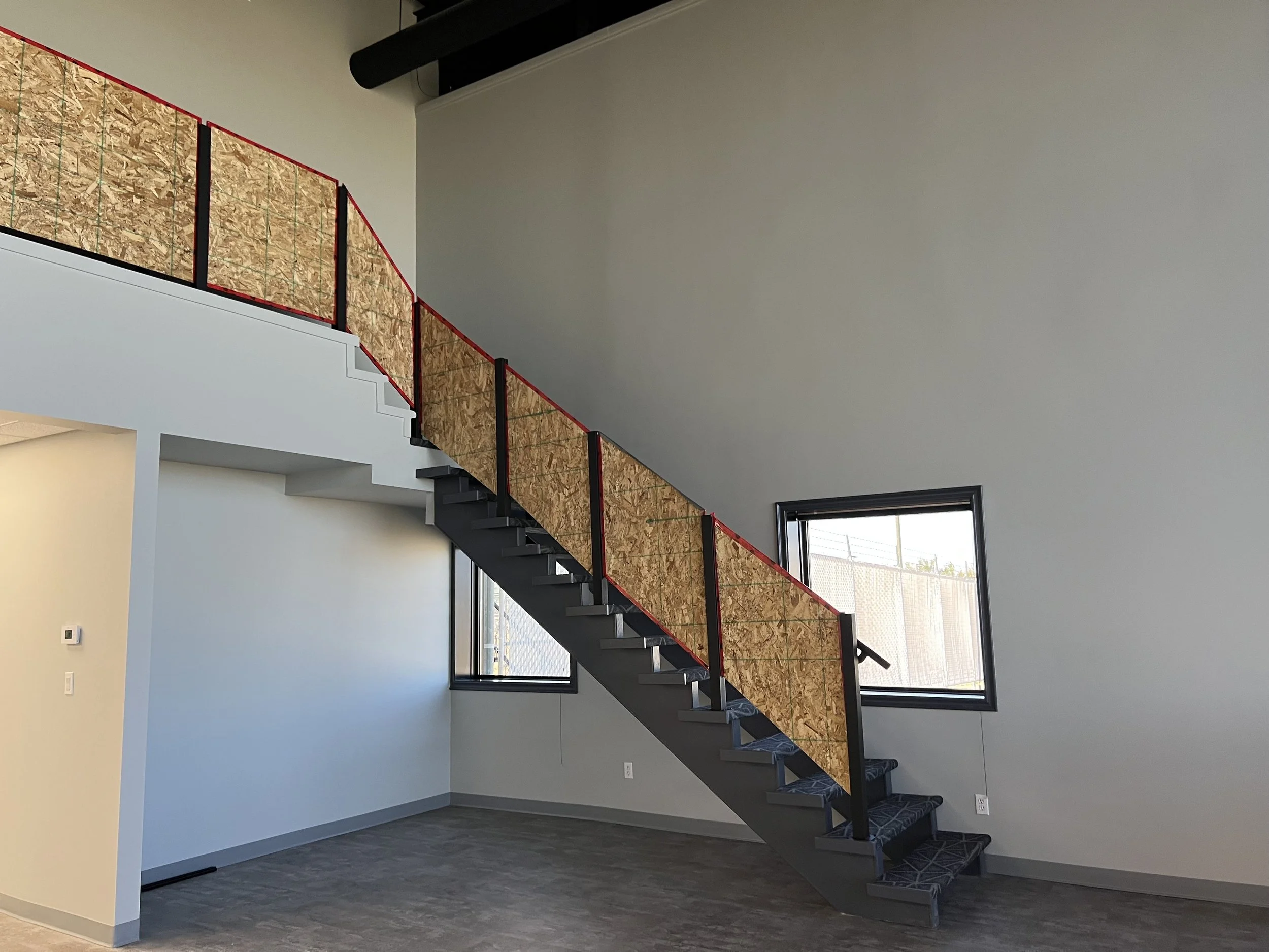 Interior view of a modern staircase with black steps and a temporary wooden and red metal railing, in a room with large windows and white walls.