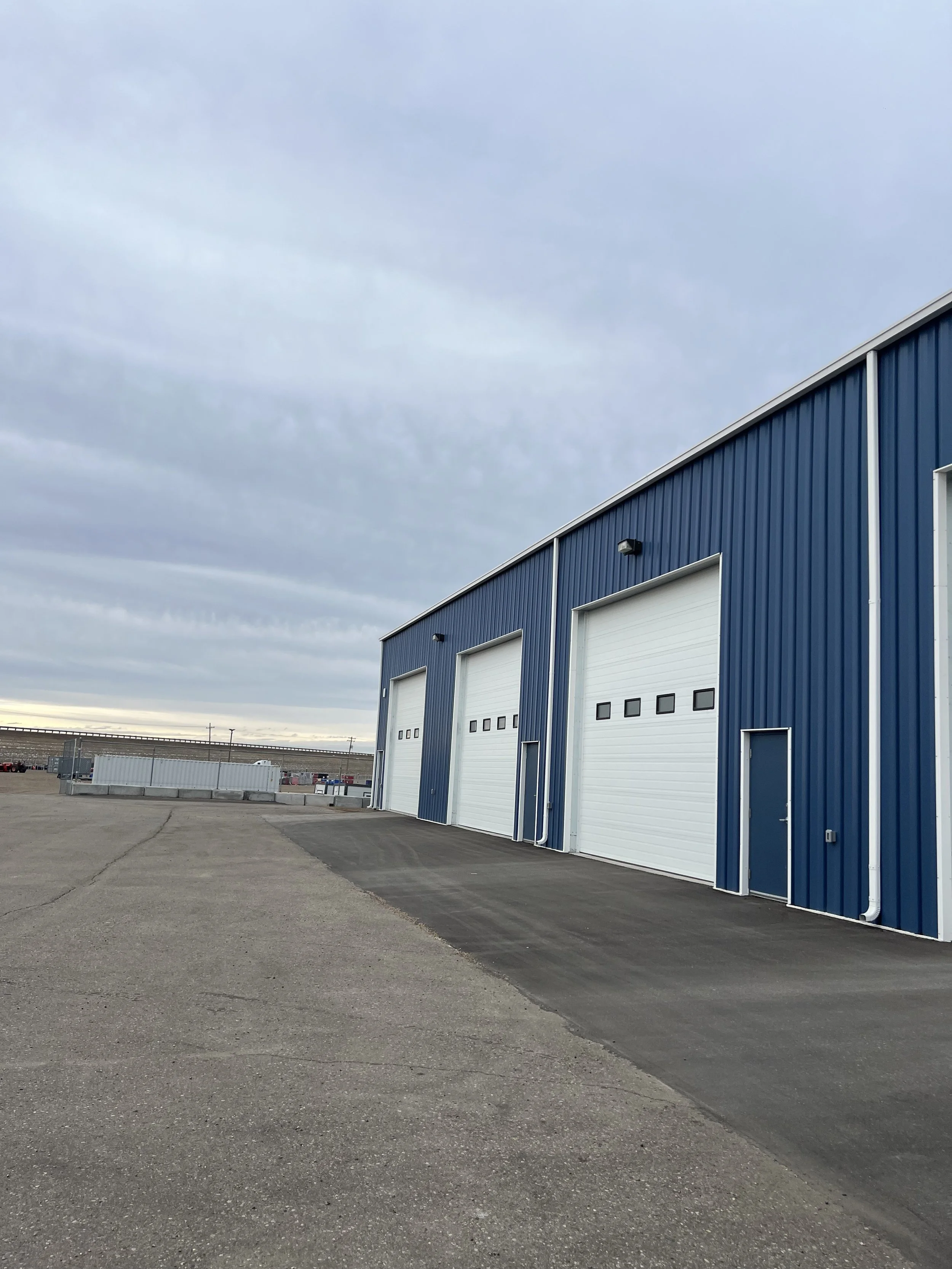 Exterior of a blue industrial building with three large white garage doors and a small pedestrian door, set against a cloudy sky.