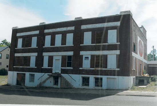 A three-story brick building with a white door and stairs leading up to it. The windows are boarded up or missing glass, and some appear to be broken. The building is situated on a street with a sidewalk.