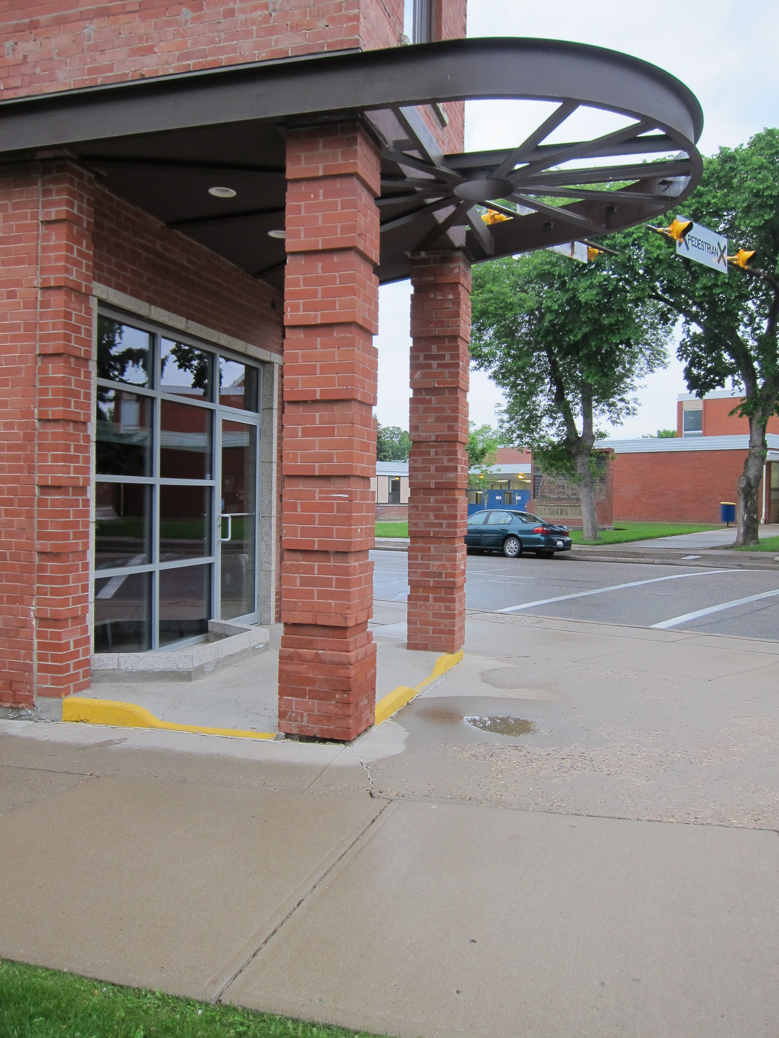 Exterior view of a brick building with large windows and a modern overhang supported by brick columns. There is a pedestrian sign and some trees in the background, along with parking lot and parked cars.
