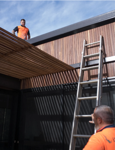 Two men working on the exterior of a building with a ladder leaning against the wall. One man is on the roof, and the other is looking up from the ground.