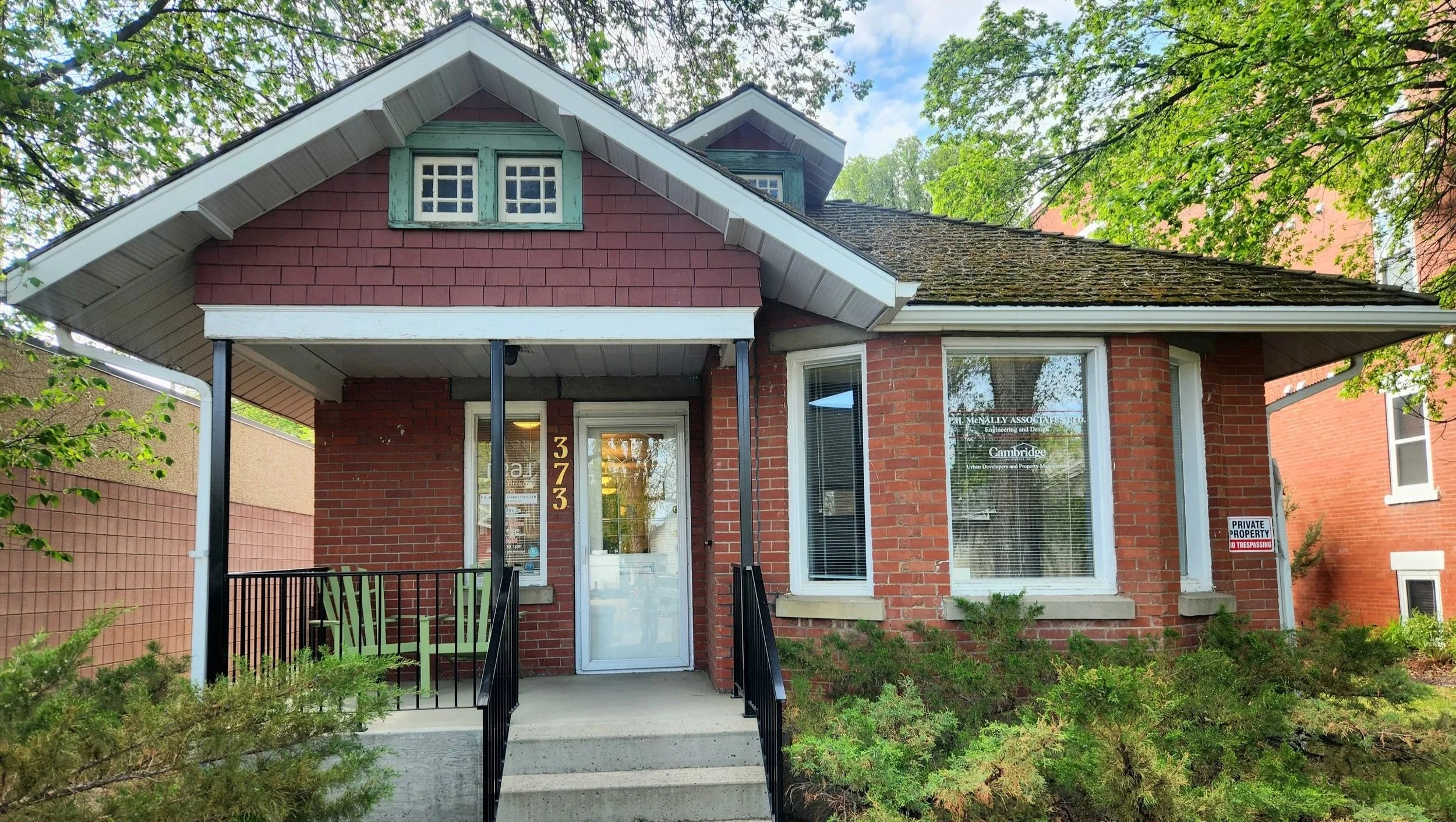 Red brick house with front porch, black railing, glass door, large window, and trees surrounding it.