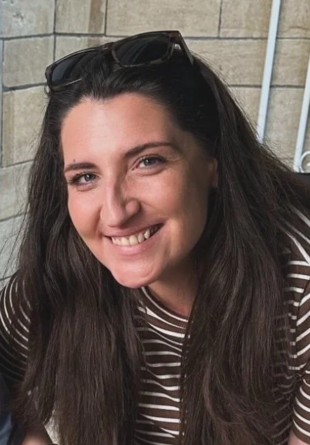 A woman with long dark hair, wearing sunglasses on her head and a striped shirt, smiling at the camera against a brick wall background.