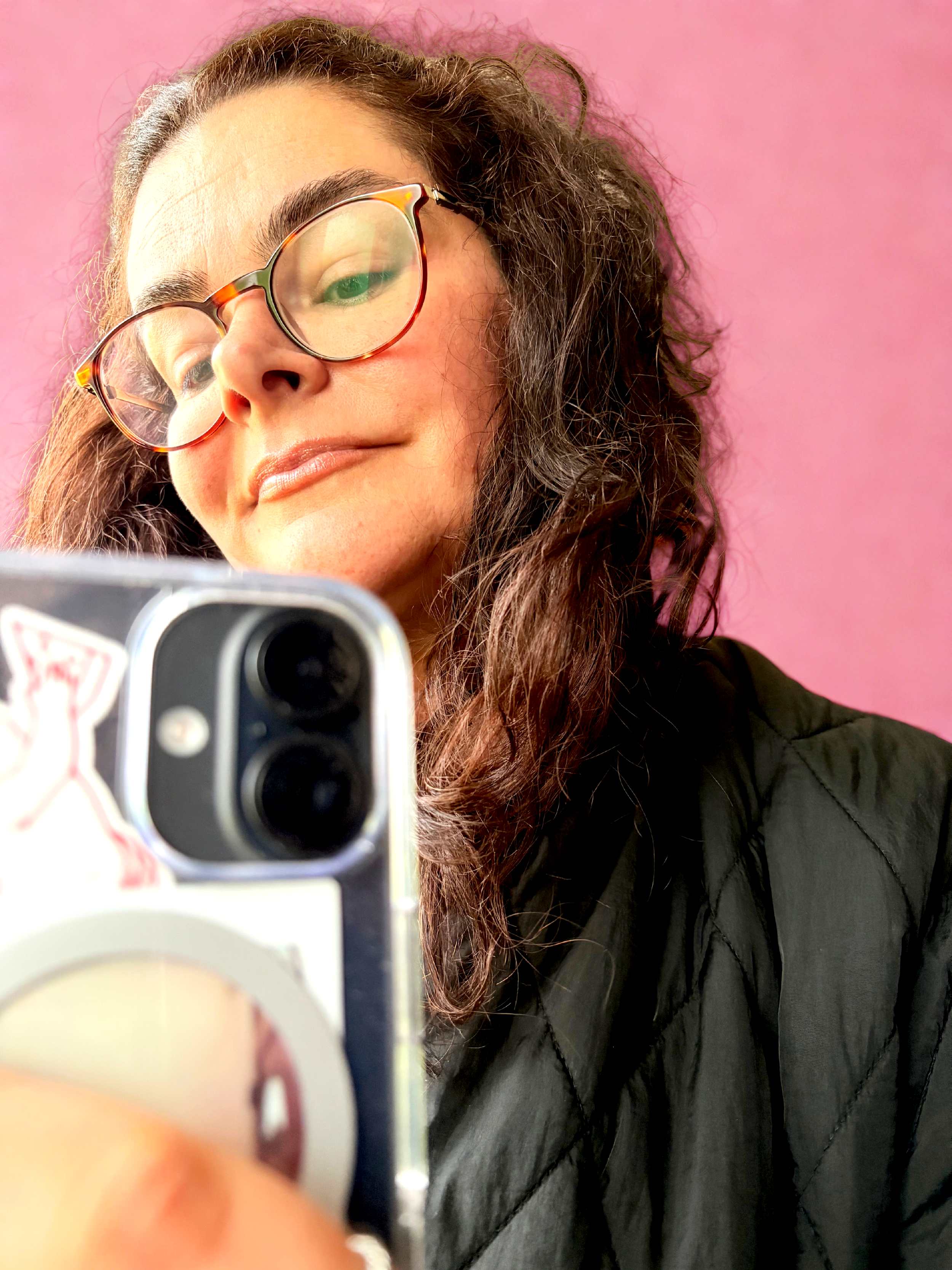 A woman with curly brown hair, glasses, and wearing a black jacket, takes a selfie with a smartphone against a pink background.