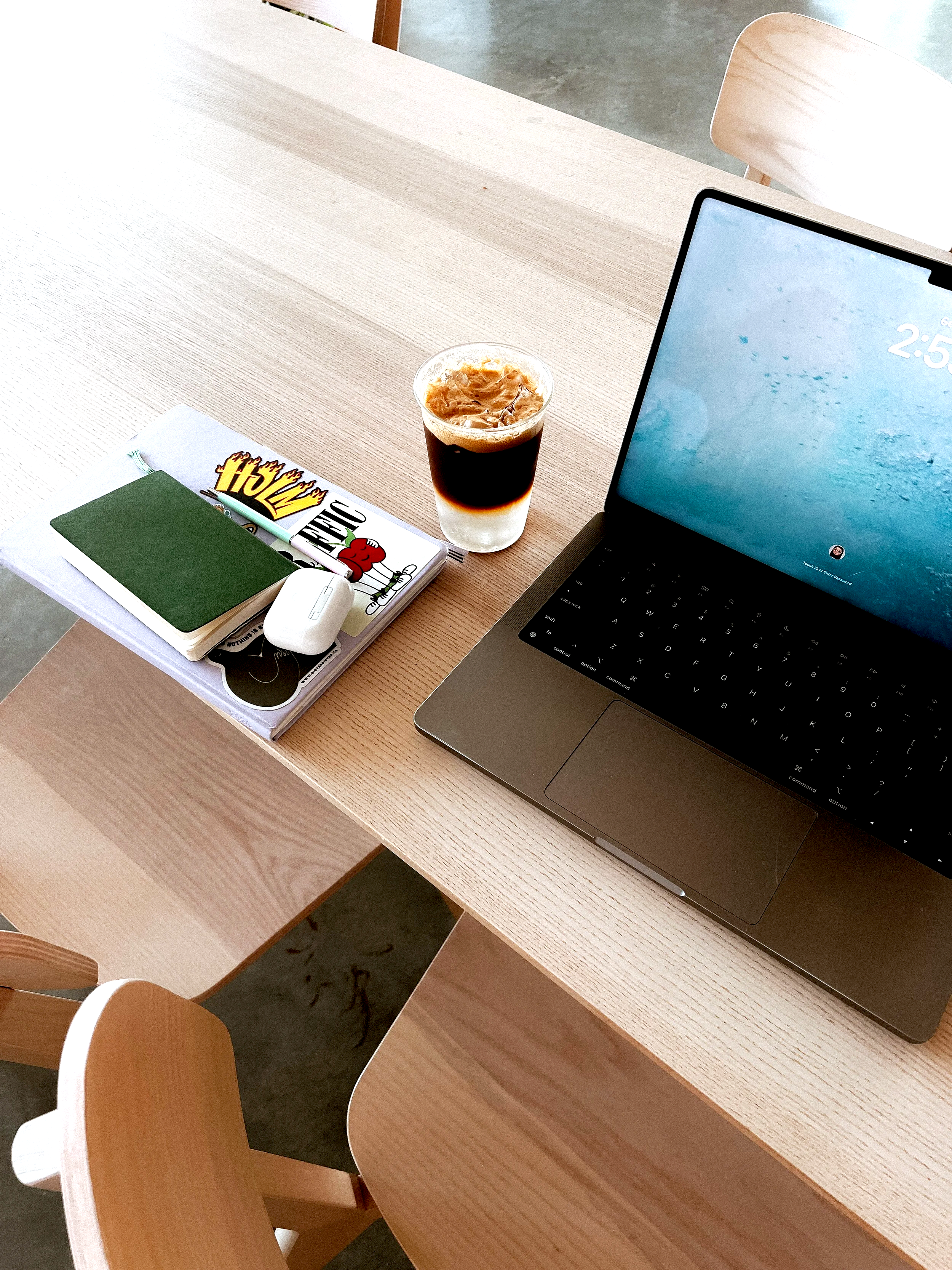 A light-colored wooden table with a laptop, a plastic cup of iced coffee with cream, a green notebook, and a white computer mouse. Two wooden chairs are visible around the table.