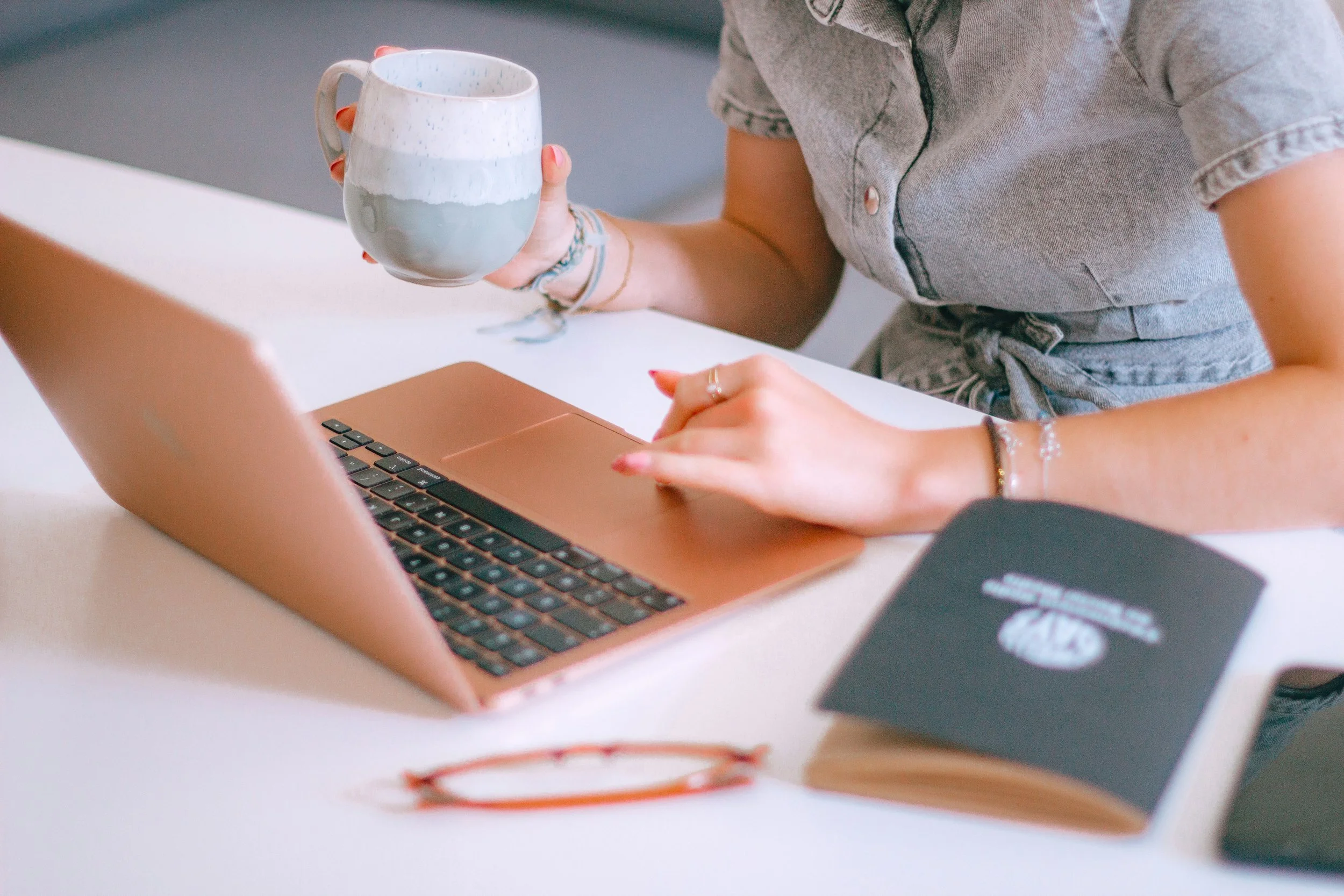 Person holding a ceramic mug sitting at a desk with a copper-colored laptop, notebook, and glasses, wearing casual clothing.