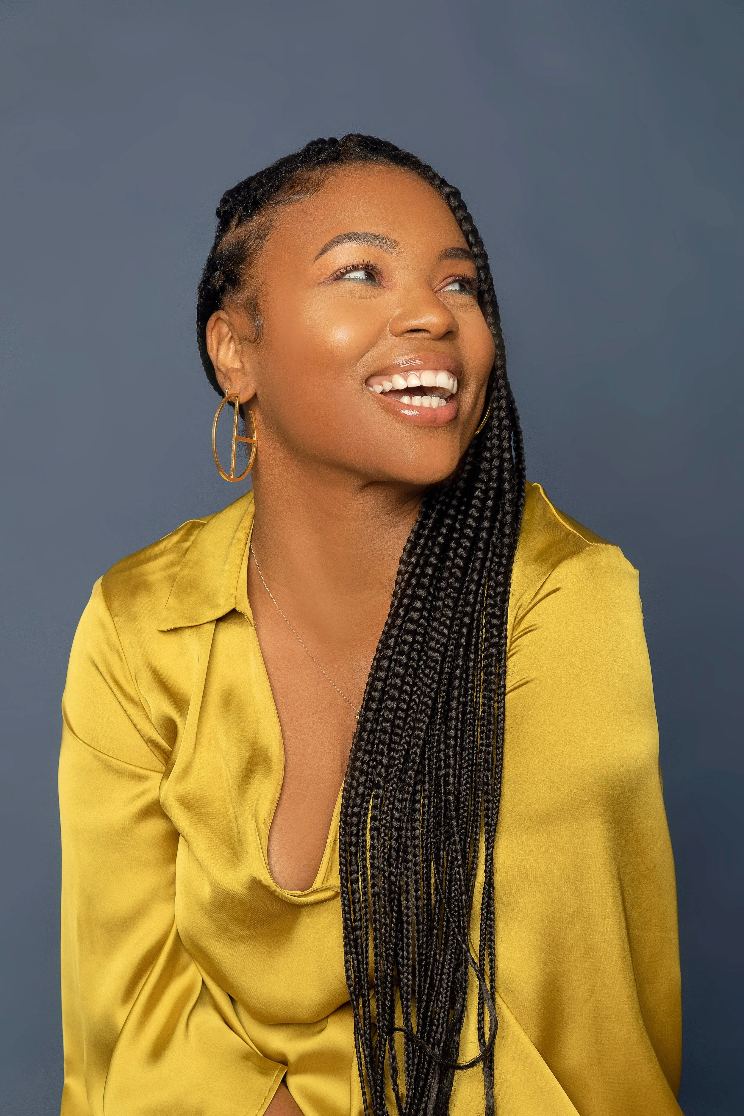 Portrait of a woman with braided hair, wearing gold hoop earrings and a yellow satin shirt, smiling while looking slightly upward against a plain gray background.