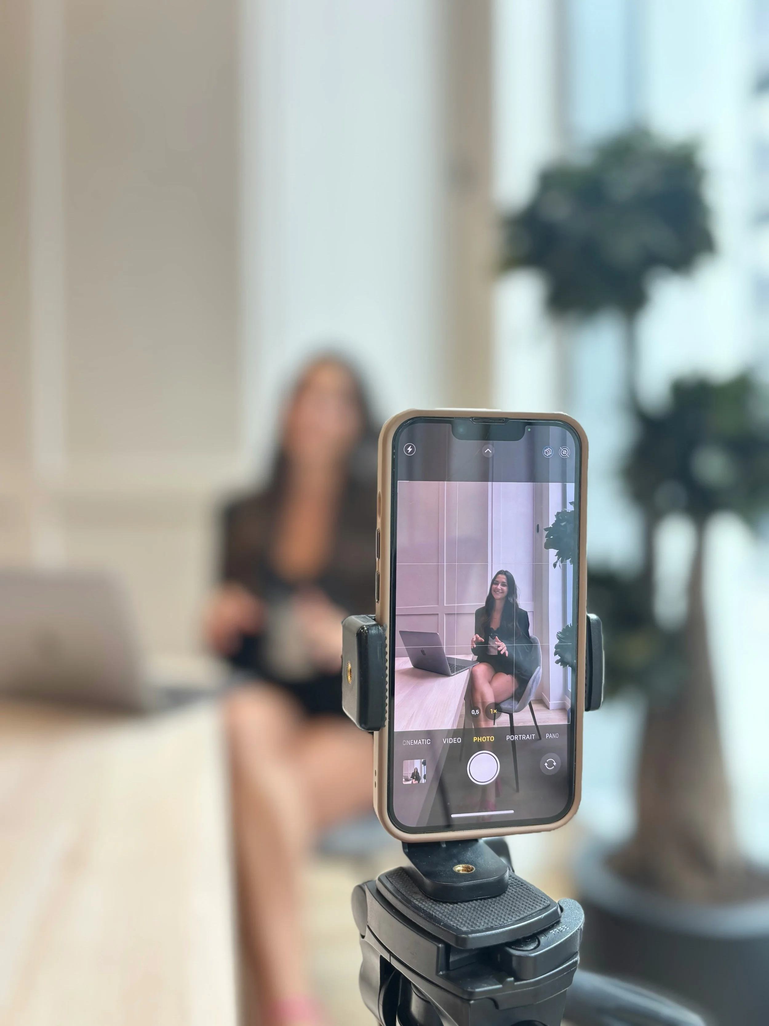 A person taking a photo or video of a woman using a smartphone mounted on a tripod or stabilizer. The woman is sitting at a table with a laptop, smiling and holding a camera or phone.