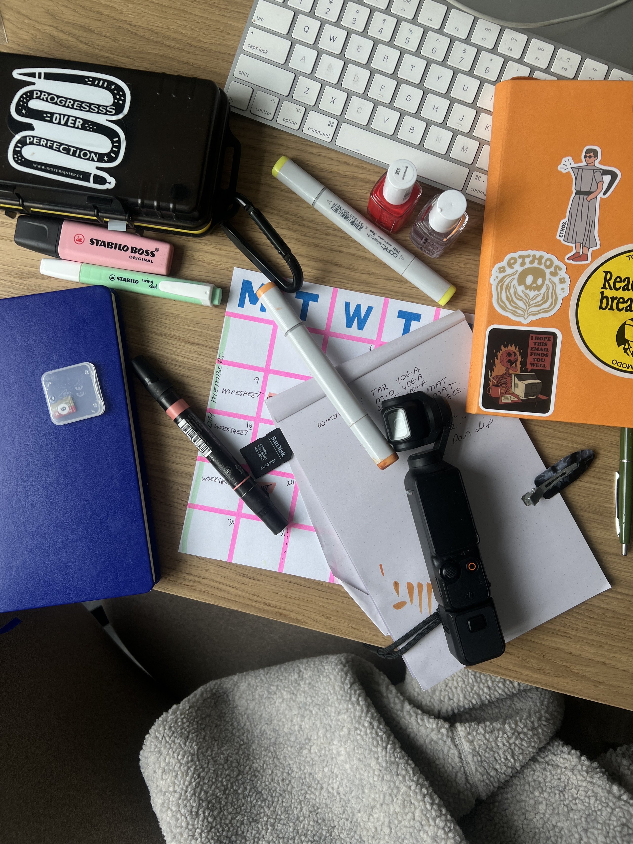 A cluttered desk with a white keyboard, various pens, highlighters, nail polish bottles, papers, a notebook with stickers, and a pair of gray fleece pants.