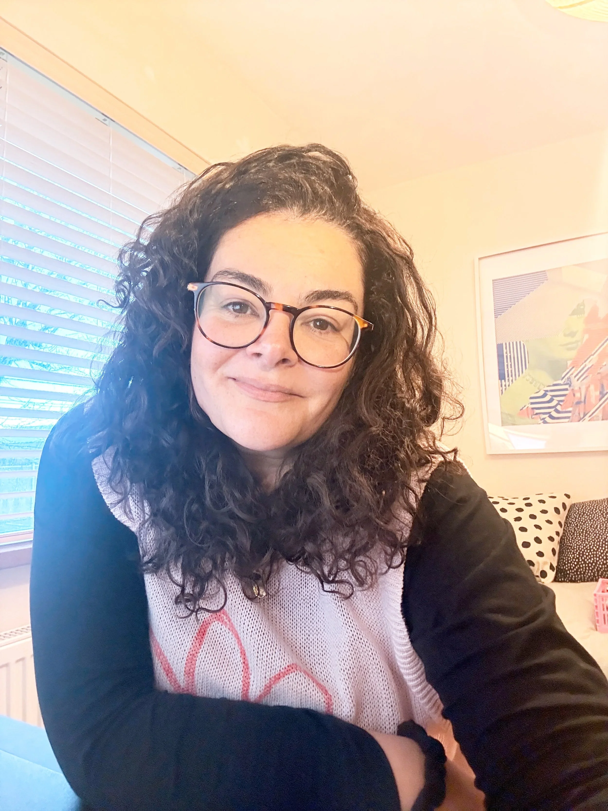 A woman with curly hair and glasses sitting indoors near a window with blinds, smiling at the camera, with a framed picture on the wall behind her.