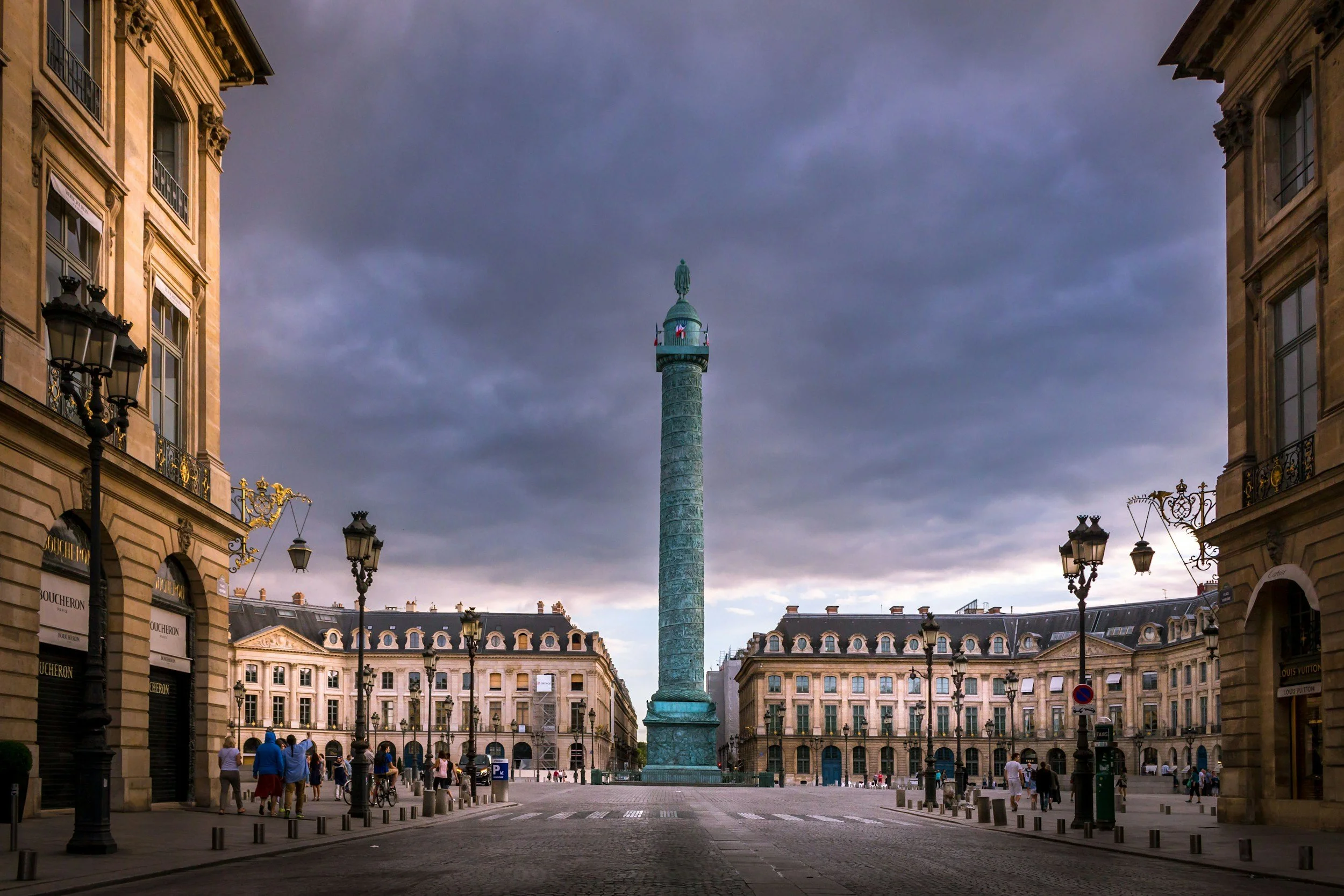 A historic city square in Paris during cloudy weather, with impressive architecture on all sides and a tall statue on a column in the center.
