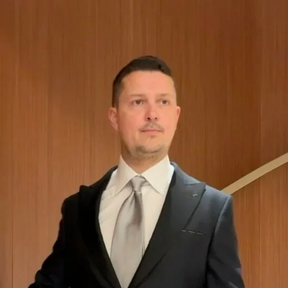 A man in a suit with a white shirt and gray tie standing in front of a wooden wall.