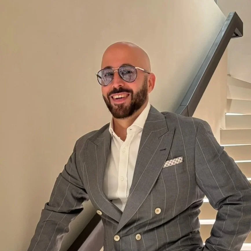 A man with a shaved head, beard, and glasses wearing a gray pinstripe suit and white shirt, standing on a staircase and smiling.