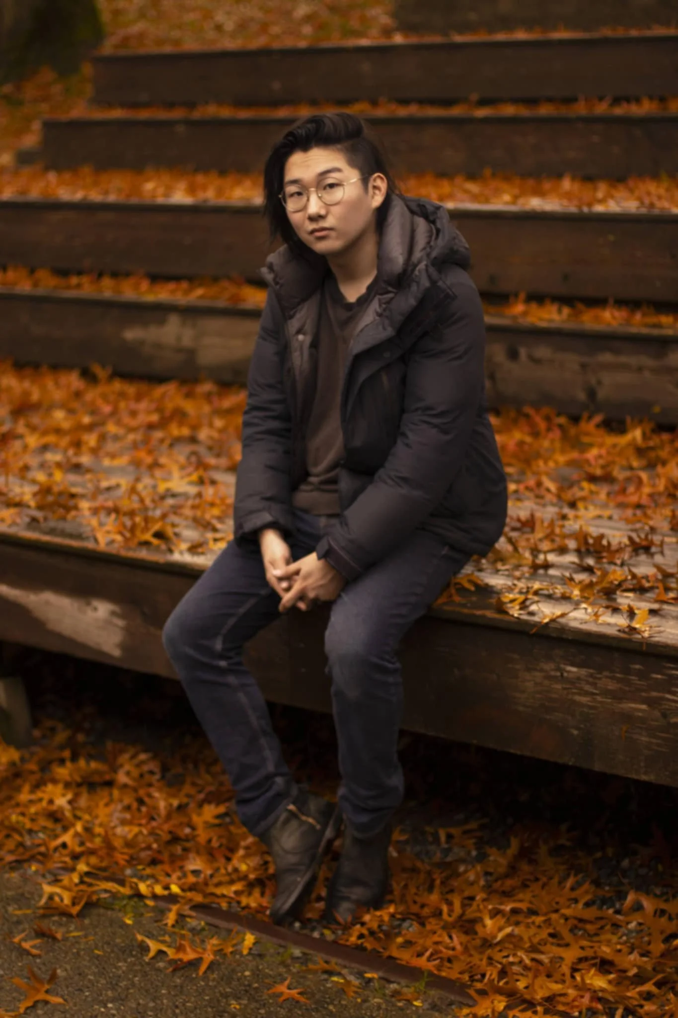 A young man with black hair and glasses sitting on a wooden bench covered in fallen autumn leaves. He is wearing a dark jacket, gray pants, and boots, with an autumn park setting in the background.