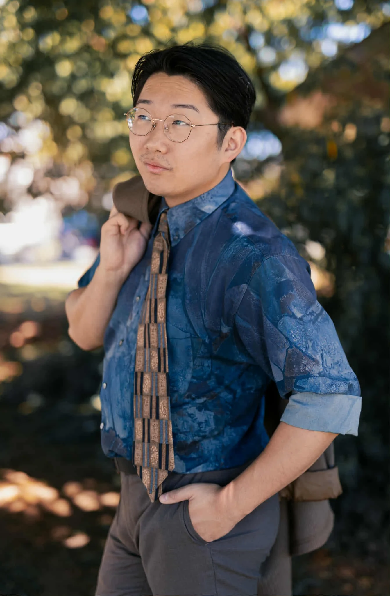 A man wearing glasses, a blue patterned shirt, and a brown tie standing outdoors in front of blurred trees with autumn foliage, holding a brown jacket over his shoulder with one hand and resting his other hand on his hip.