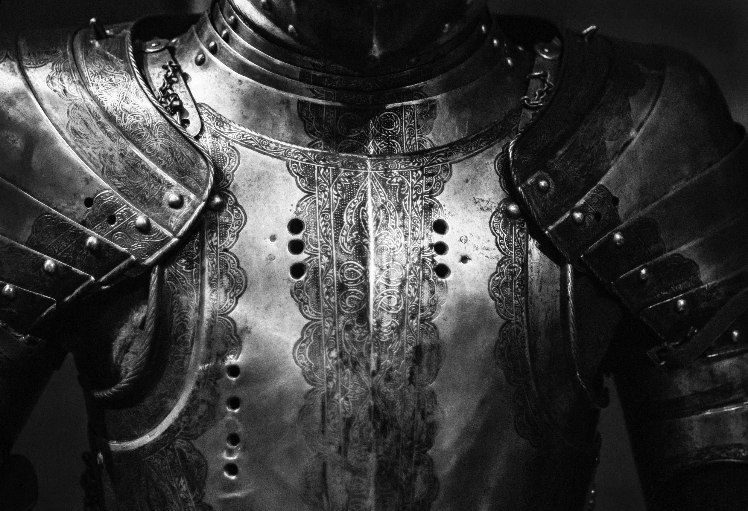 Close-up black and white photo of a suit of medieval knight's armor, showing detailed engravings, rivets, and chainmail.