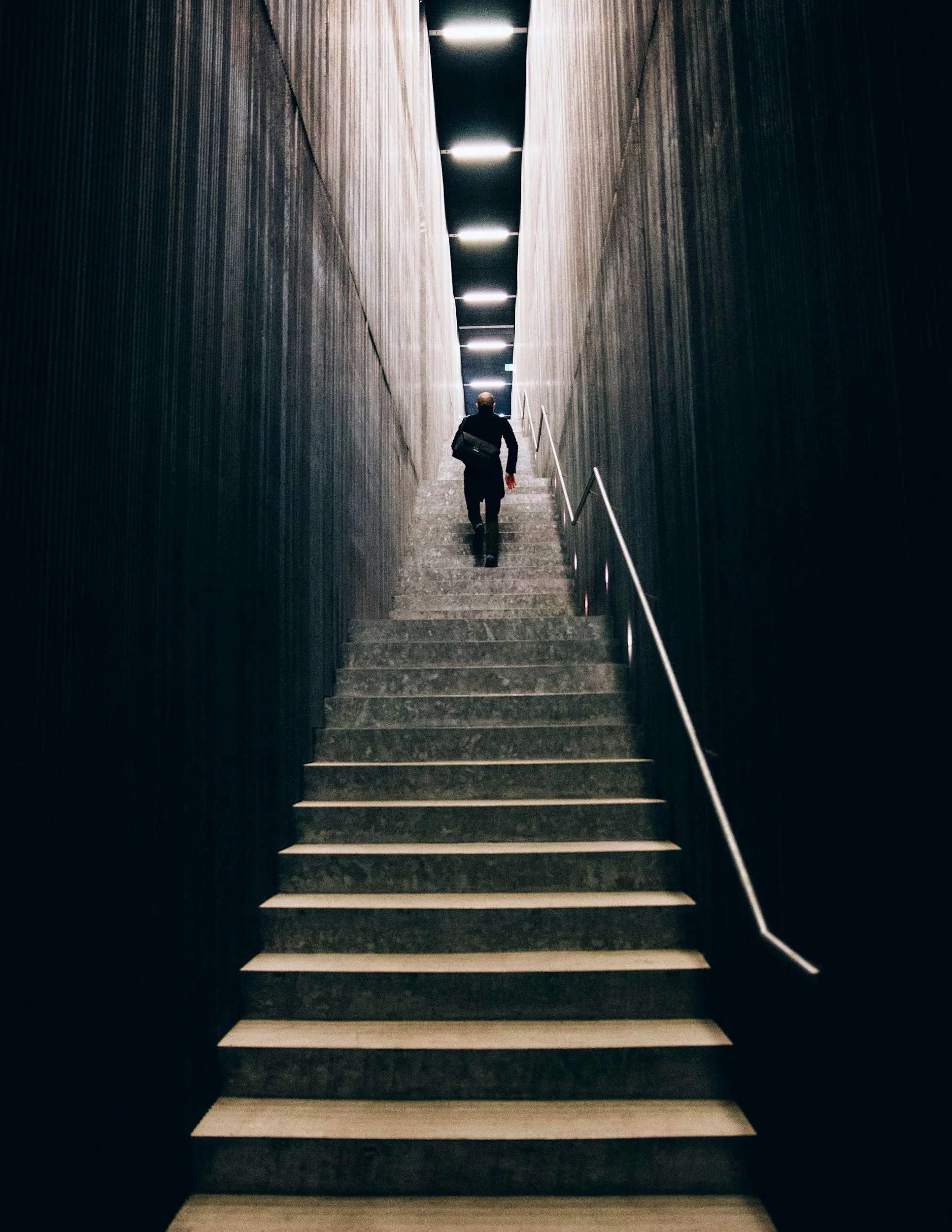 A person with a black backpack ascending a dark staircase with dark walls and bright overhead lights.
