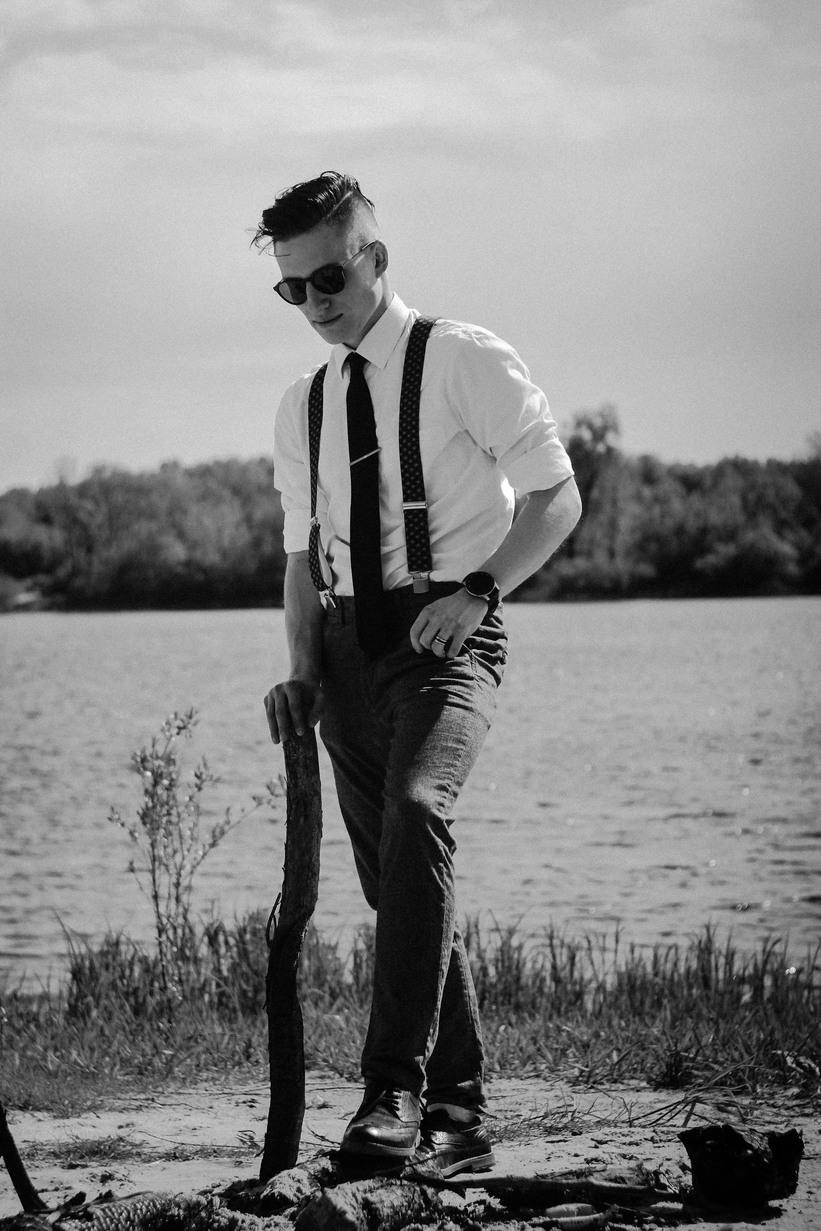 A young man wearing sunglasses, a white shirt, tie, suspenders, and dark pants standing outdoors on a beach, holding a stick or pole, with a body of water and trees in the background in black and white.