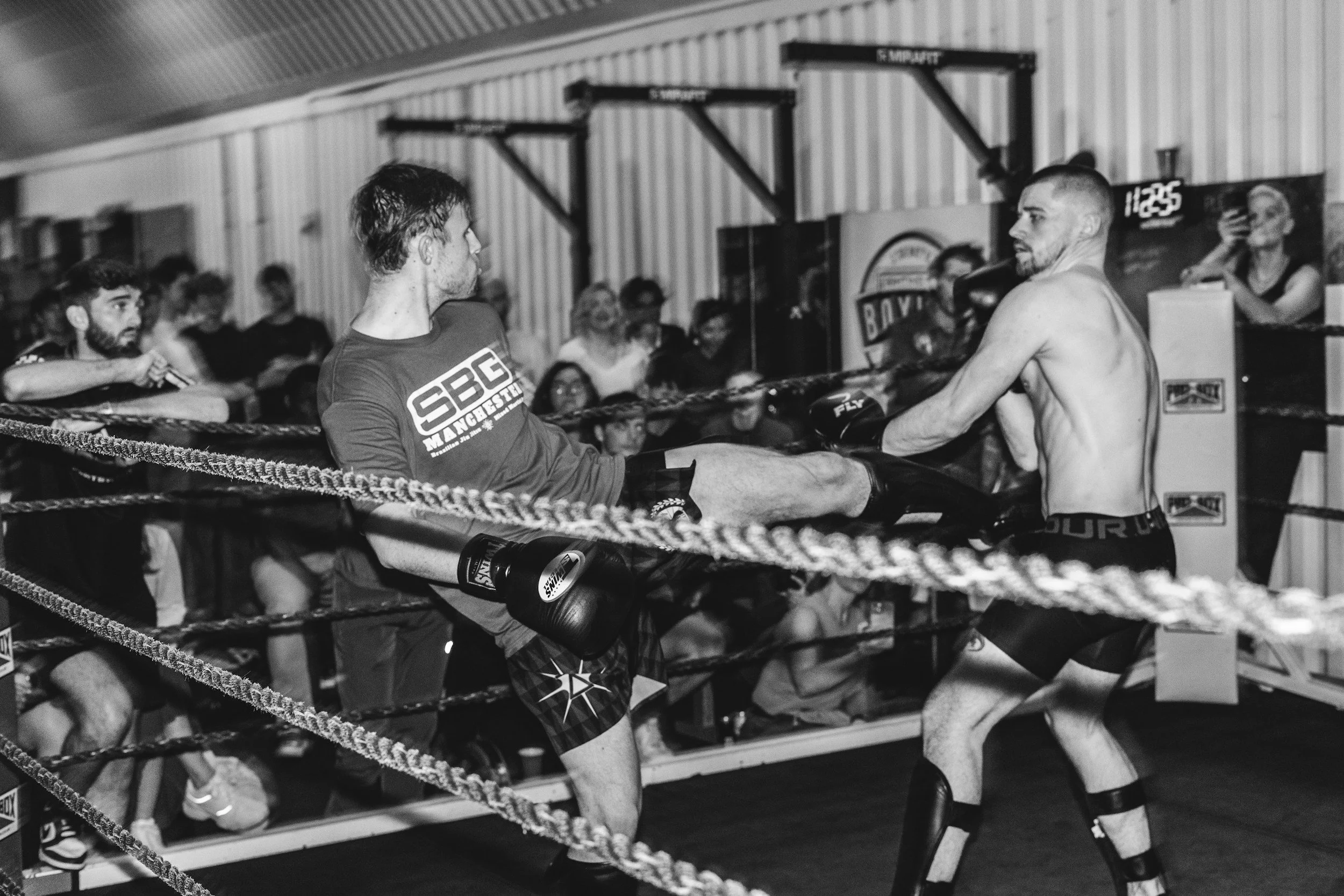 Two shirtless male fighters sparring in a boxing ring with ropes, fighting gloves, and athletic shorts, surrounded by an audience in a gym or training facility.