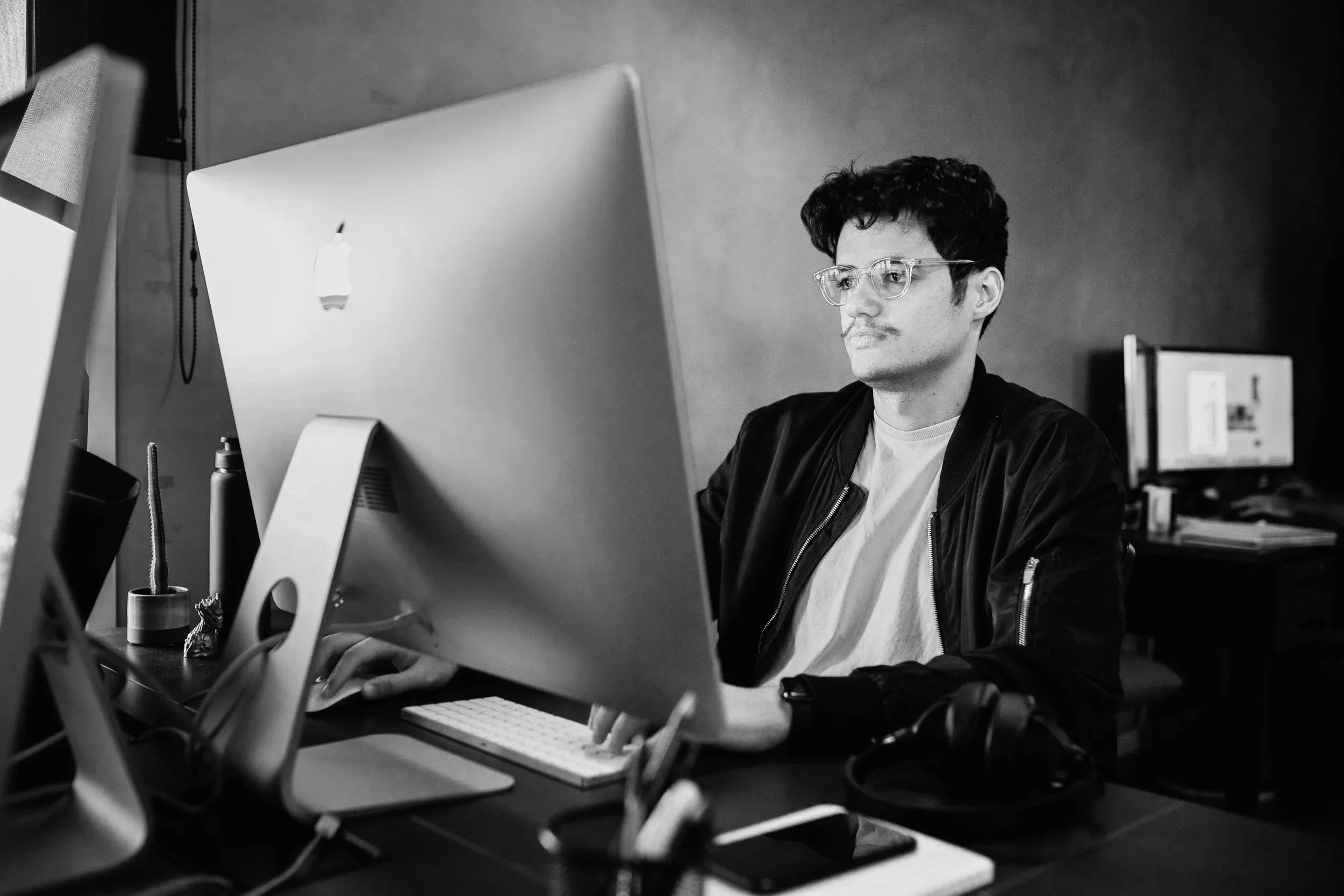 A young man with curly hair and glasses working at a computer in a modern office environment, with another monitor visible in the background.
