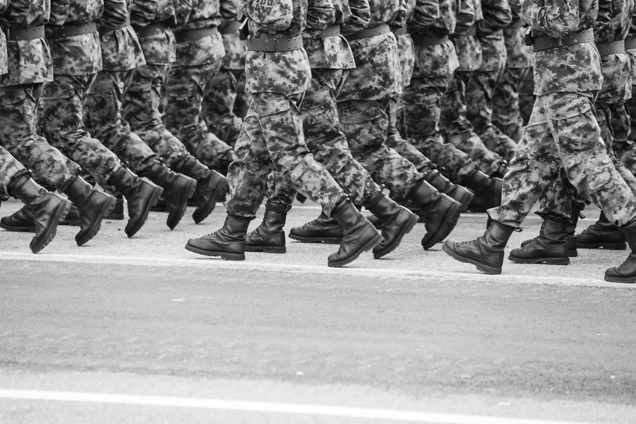 Black and white photo of soldiers in camouflage uniforms marching in formation, showing their legs and boots.