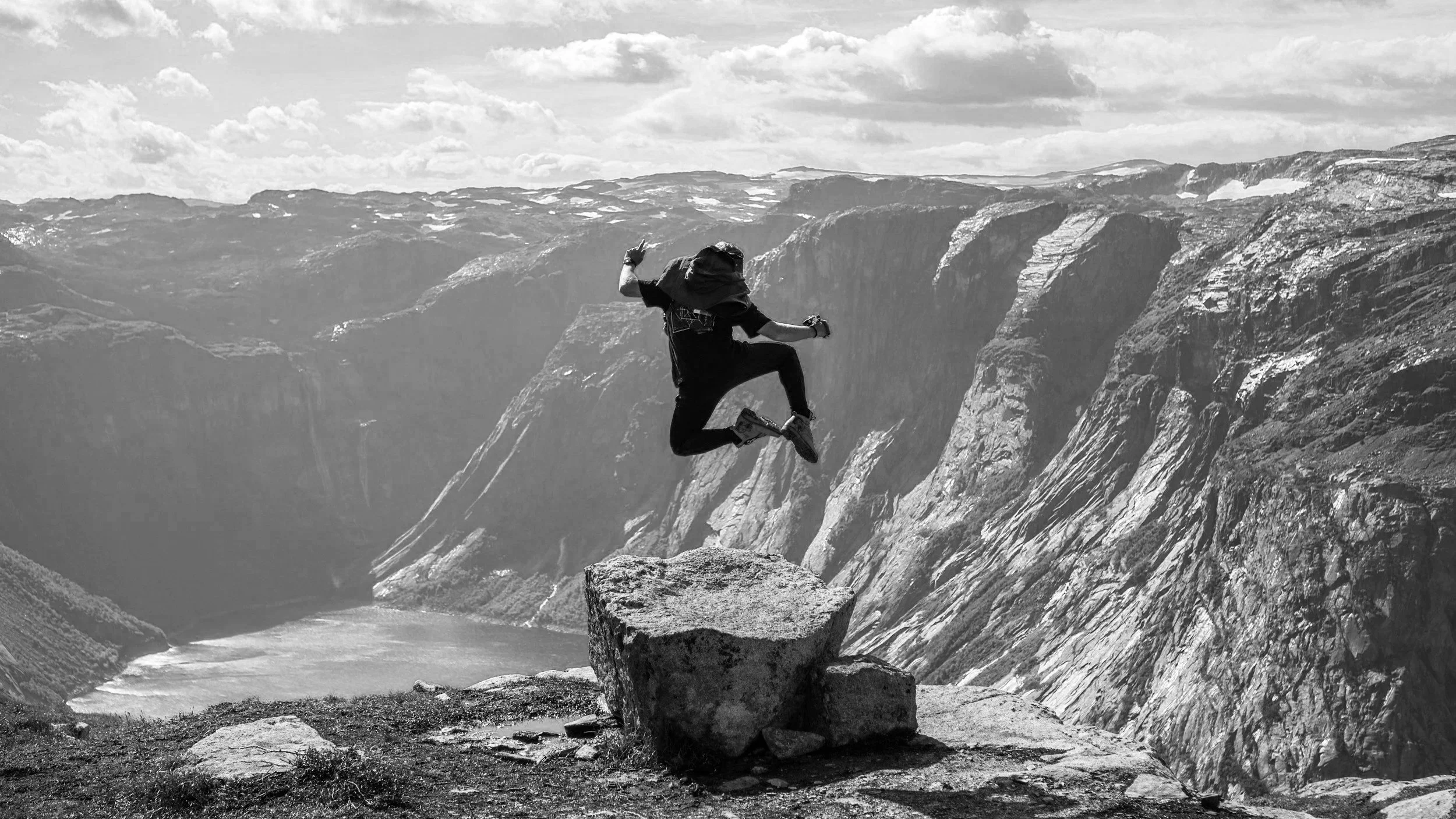 A person wearing a hat and dark clothing jumping off a large rock in a mountainous landscape with steep cliffs and a river below.
