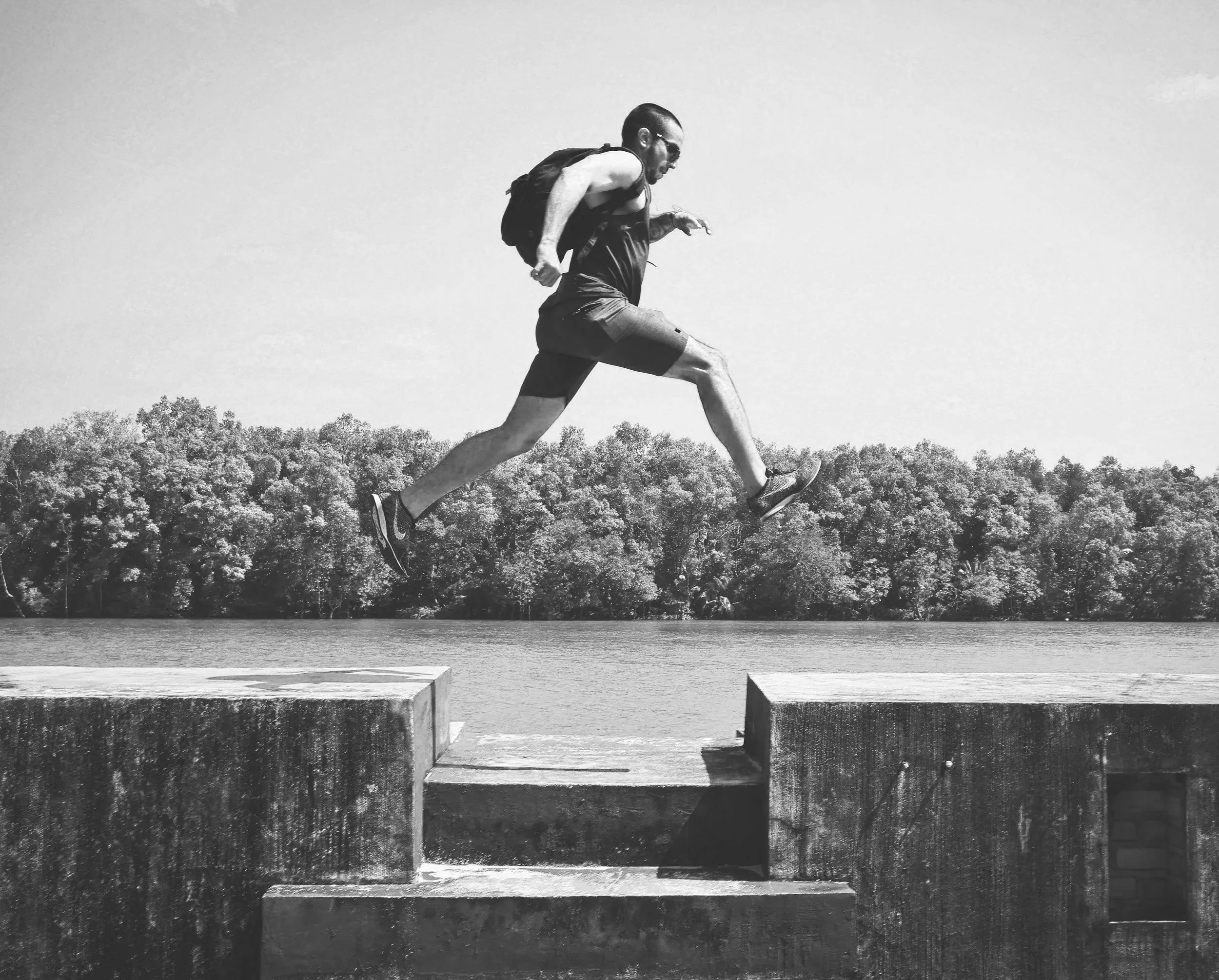 A man running with a backpack in a park with trees and water in the background, seen in black and white.