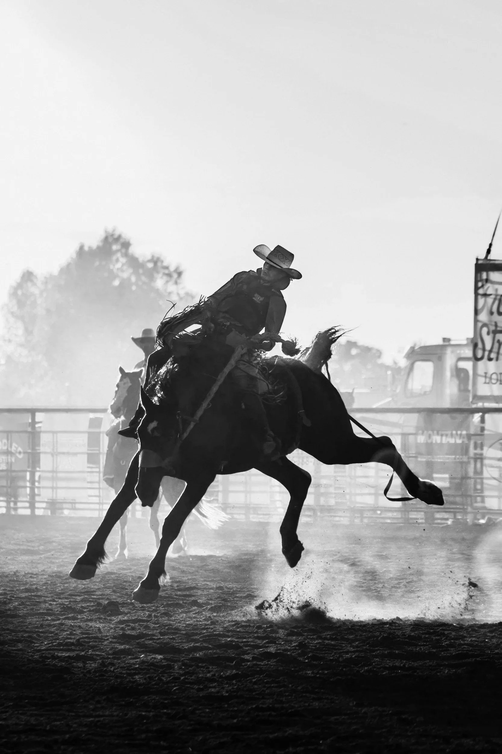 A cowboy riding a bucking horse at a rodeo, with dust in the air and a sign in the background.