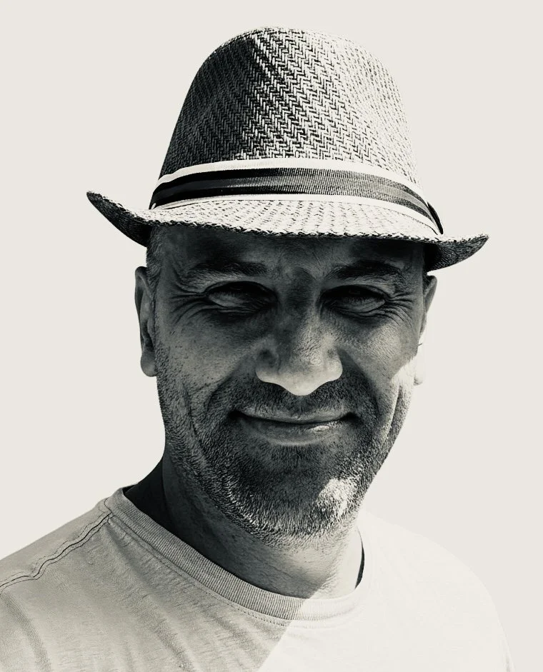 Smiling man wearing a patterned fedora hat and a light-colored T-shirt, with a beard and mustache, looking at the camera.