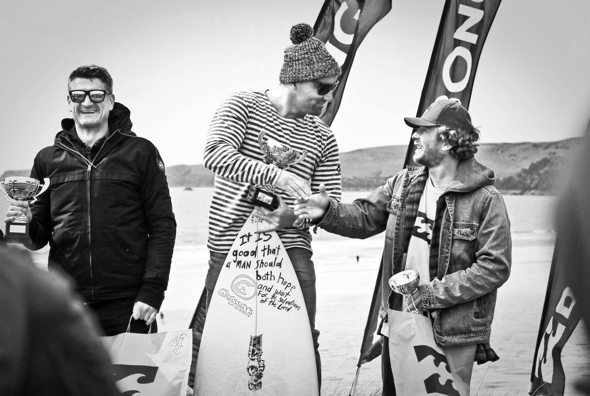 Three men at a beach holding trophies, with banners and surfboards in the background. One man is smiling, another is receiving a trophy, and the third is handing him the trophy, wearing casual beach clothing and sunglasses.