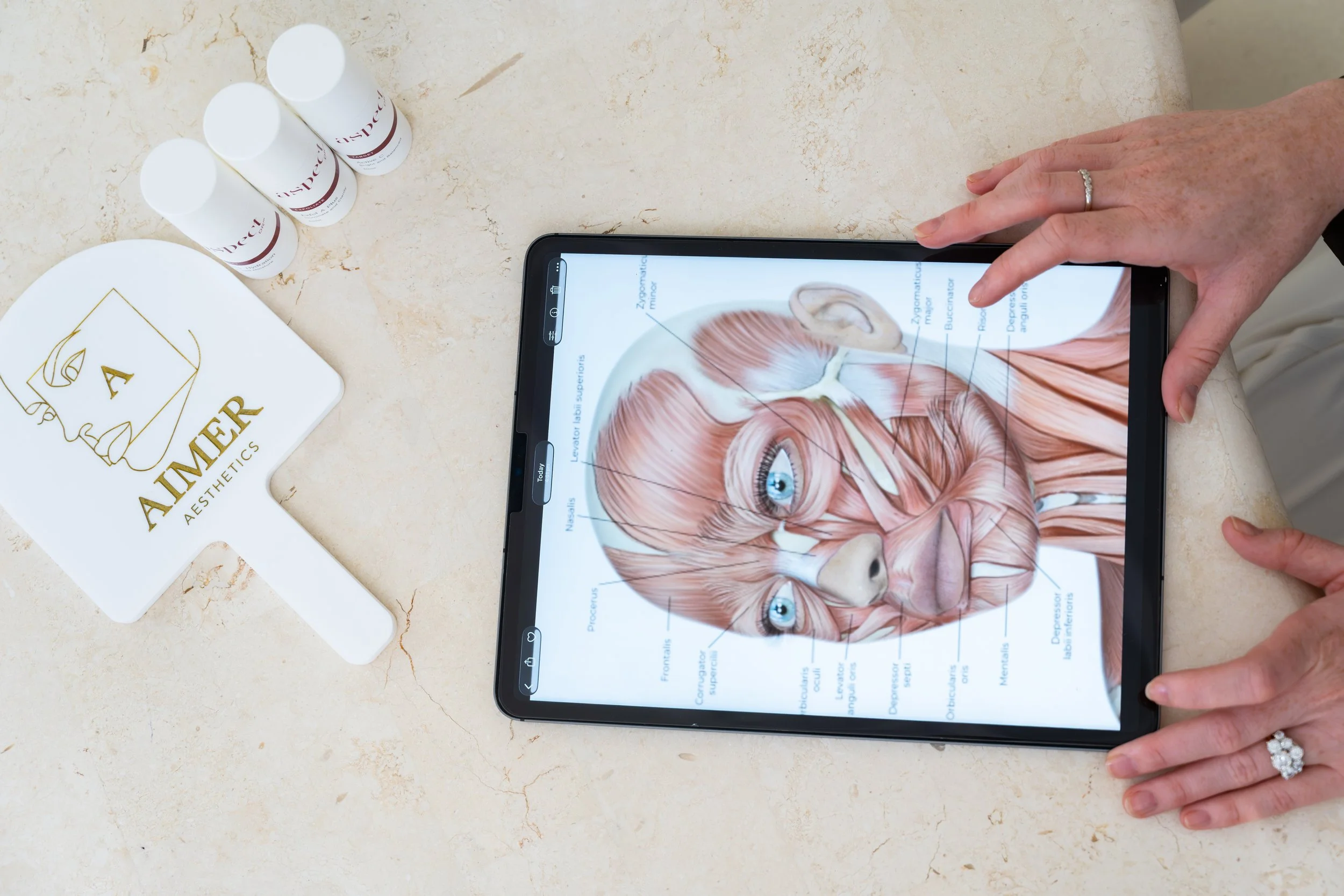 A person with a wedding ring using a tablet showing a detailed diagram of the human head with muscles and nerves labeled. On the table, there are three white bottles labeled 'Deca.' Also present is a white cloth with a gold logo and text that reads 'L AIMER AESTHETICS.'