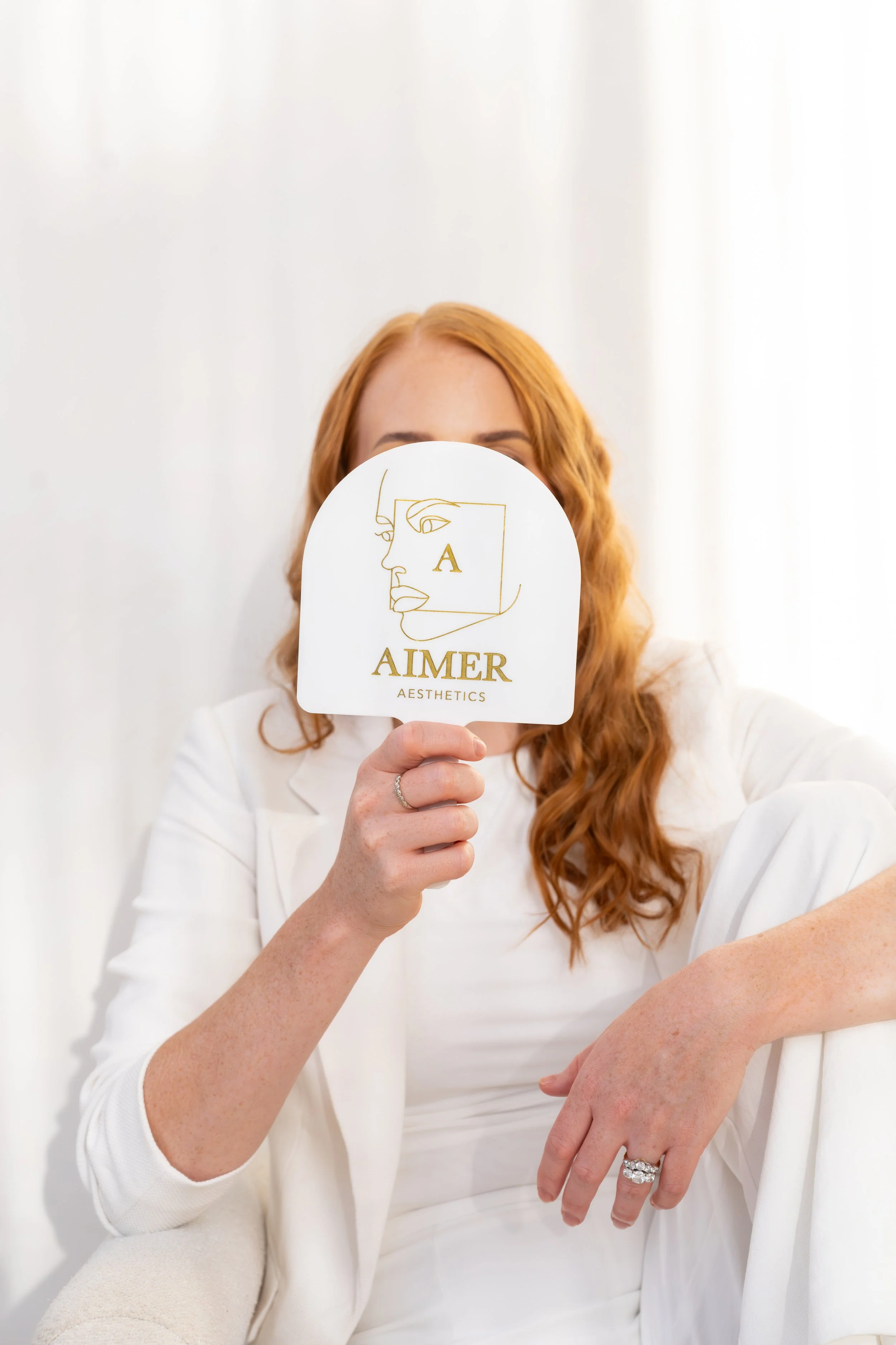 Woman with long red hair holding a round sign that reads "Aimer Aesthetics" in front of her face, wearing a white outfit, sitting on a white armchair against a white background.
