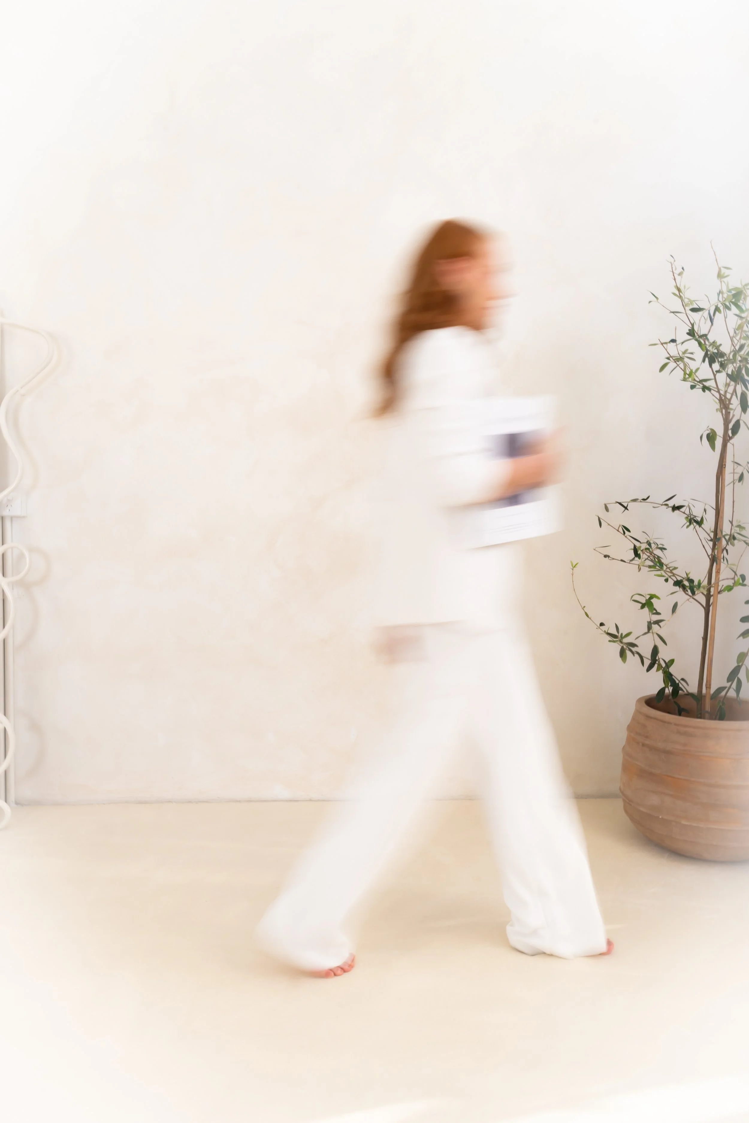 Blurry woman in white clothing practicing yoga in a minimalistic room with a potted plant.