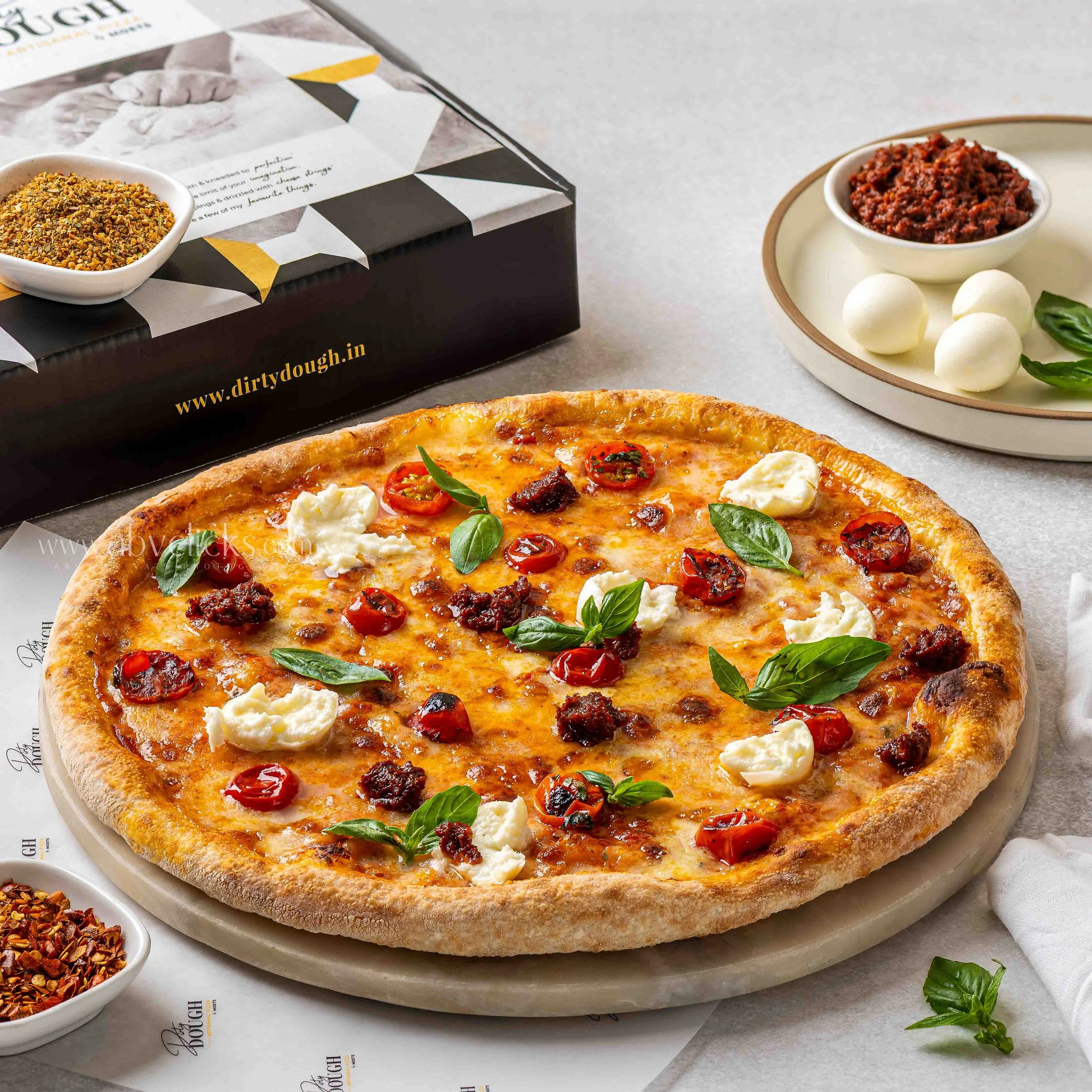 A pizza with cherry tomatoes, mozzarella, basil, and ground beef on a white surface next to a dish of red pepper flakes and a bowl of red chili paste.