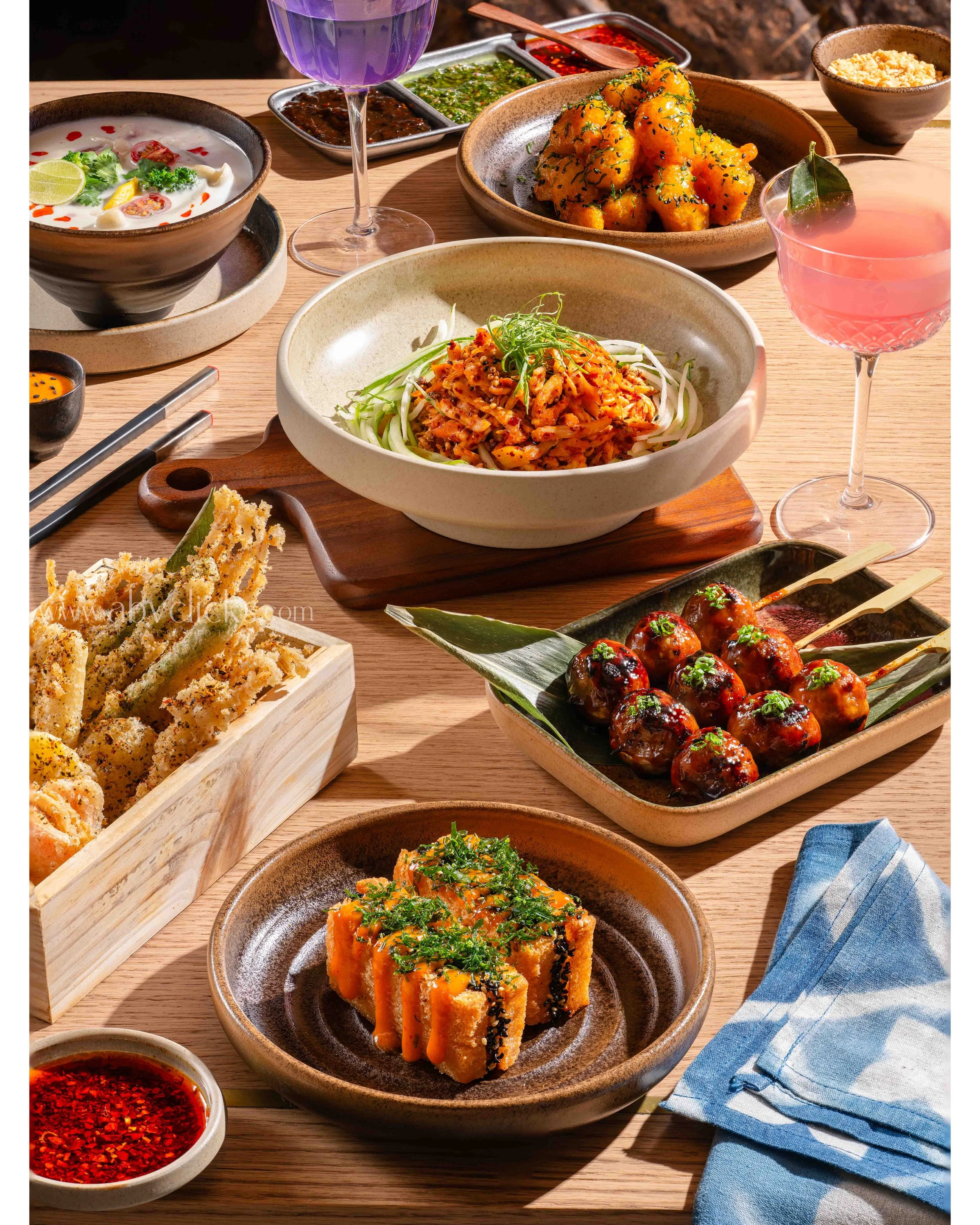 A variety of Asian dishes including dumplings, a salad, skewered meatballs, and dipping sauces, served on wooden and ceramic plates with colorful drinks and a blue checkered napkin.