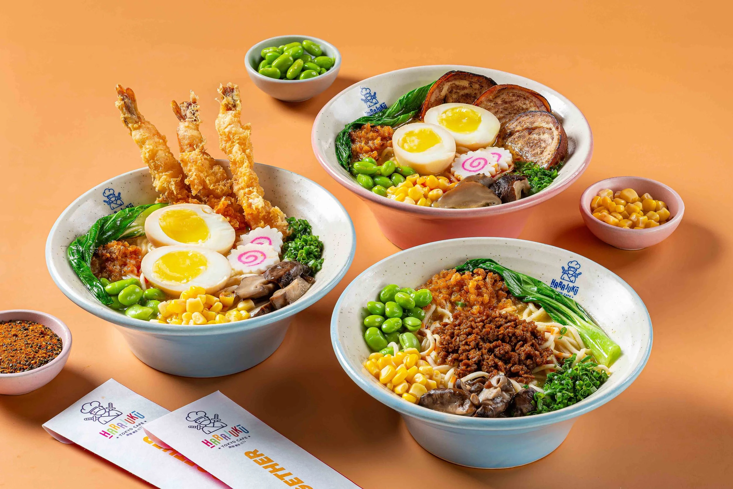 Multiple bowls of ramen with boiled eggs, green vegetables, and various toppings on a peach-colored surface. Small bowls of corn and spices are also visible.