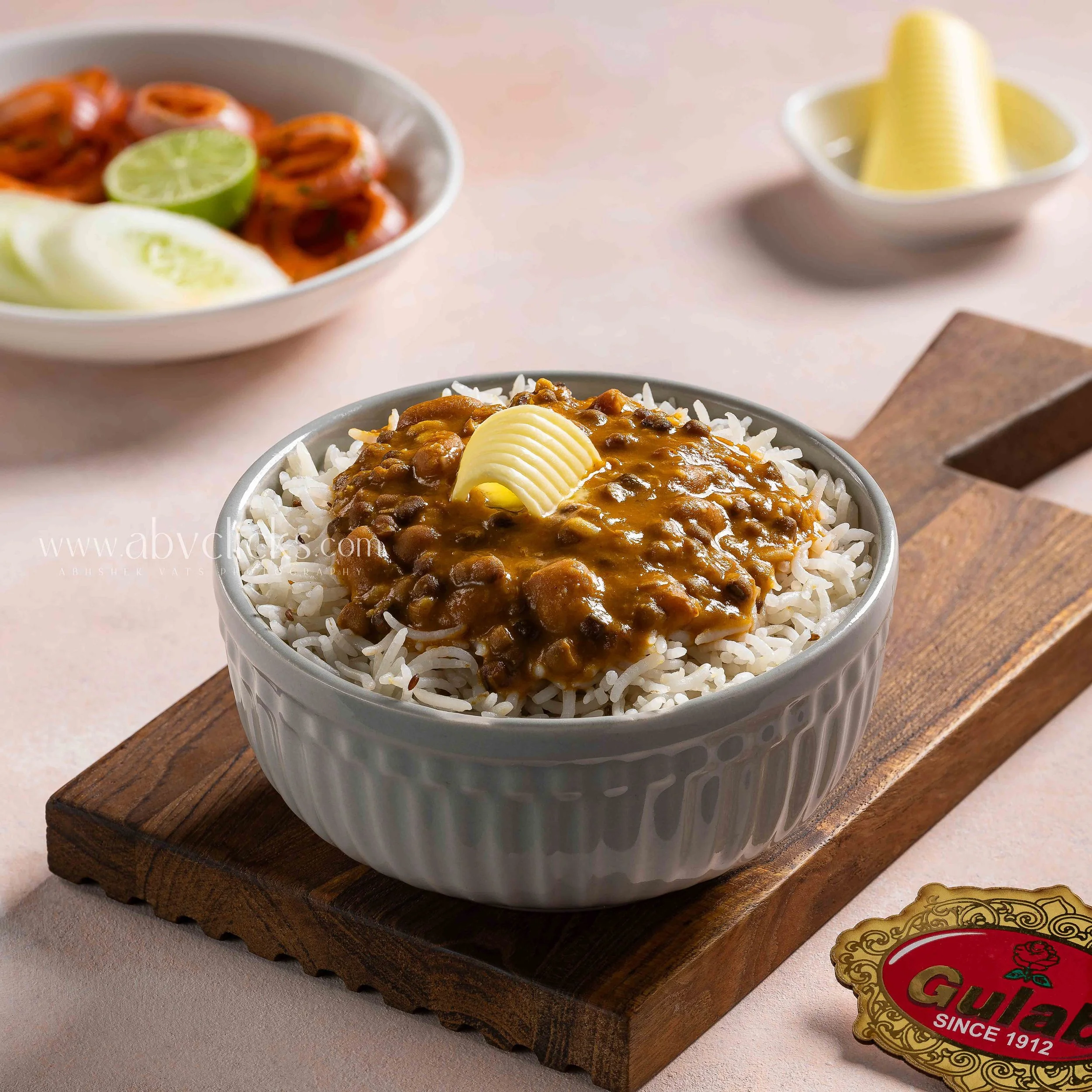 Close-up of a bowl of white rice topped with curry, garnished with a slice of butter and a quartered boiled egg, placed on a wooden serving board with a decorative gold and red label.
