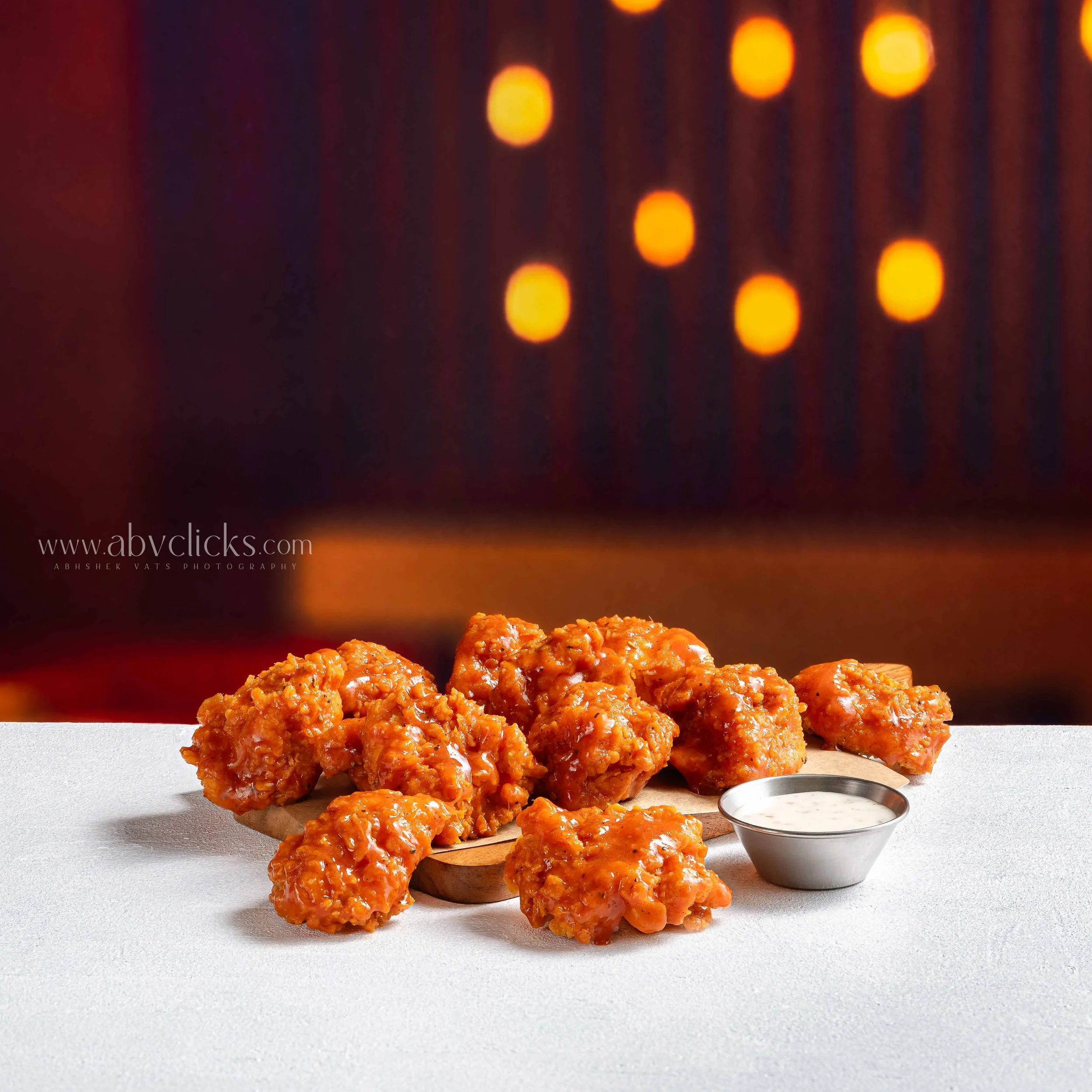 Close-up of crispy chicken pieces coated in sauce, on a white surface with a small bowl of white dipping sauce nearby. Blurred orange and yellow lights in the background.