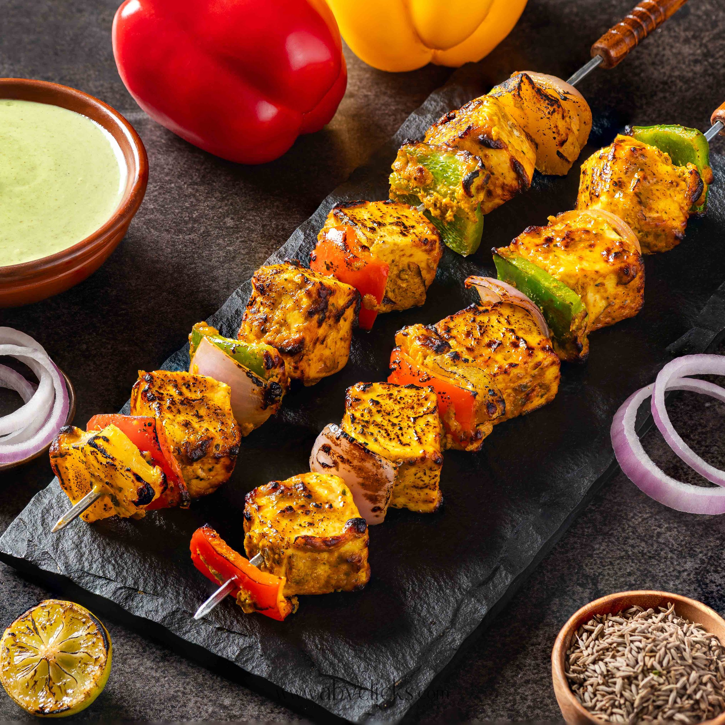 Close-up of grilled vegetable skewers with peppers, zucchini, and onions on a black slate platter, with a bowl of green sauce, grilled lemons, and a red bell pepper in the background.