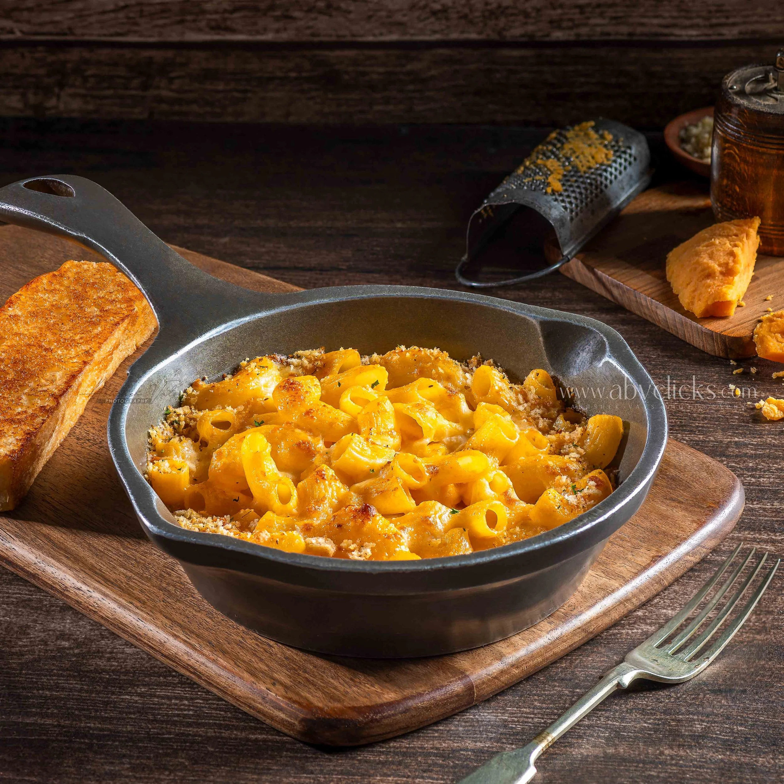 Close-up of a cast iron skillet with scrambled eggs and melted cheese, placed on a wooden cutting board with a toasted bread slice. In the background, a cheese grater, a jar of sauce or syrup, and a small bowl are visible on a dark wooden surface.