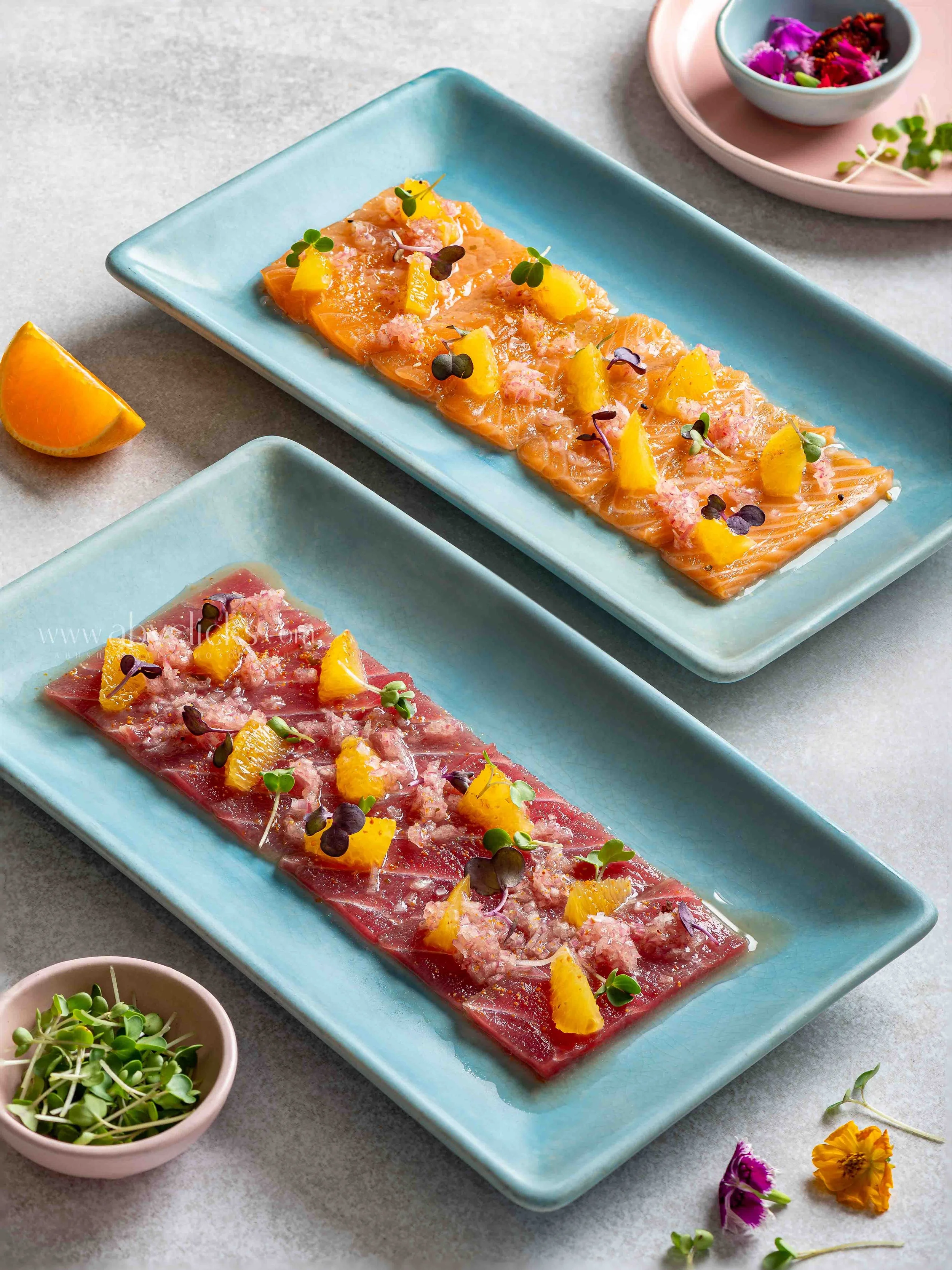 Slices of raw salmon and beef topped with pineapple chunks, microgreens, and pink coarse salt on rectangular blue plates, with small bowls of microgreens and edible flowers on a gray surface.
