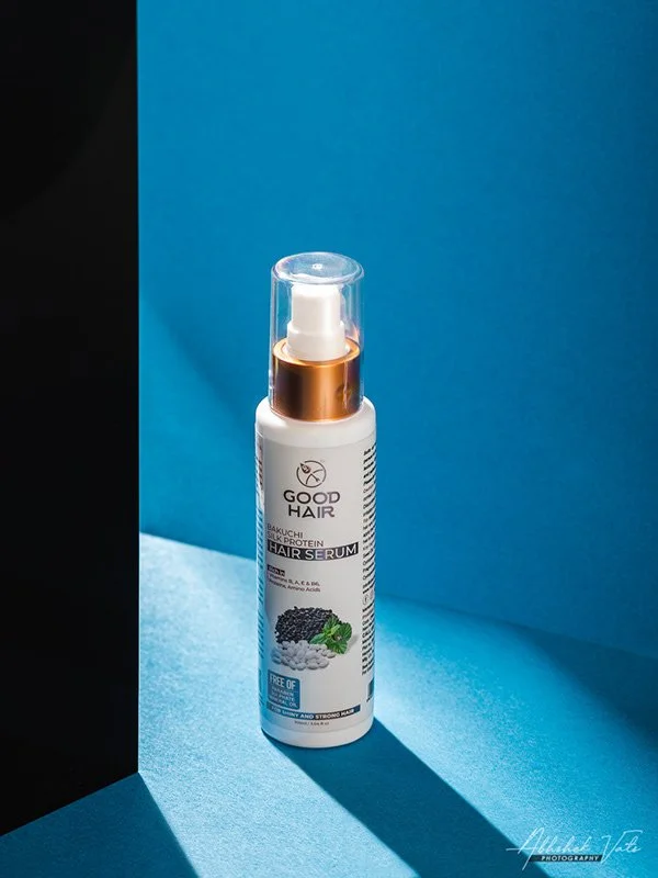 A white bottle of Good Hair hair serum with a pump dispenser, placed on a blue surface with a blue background, casting a shadow.